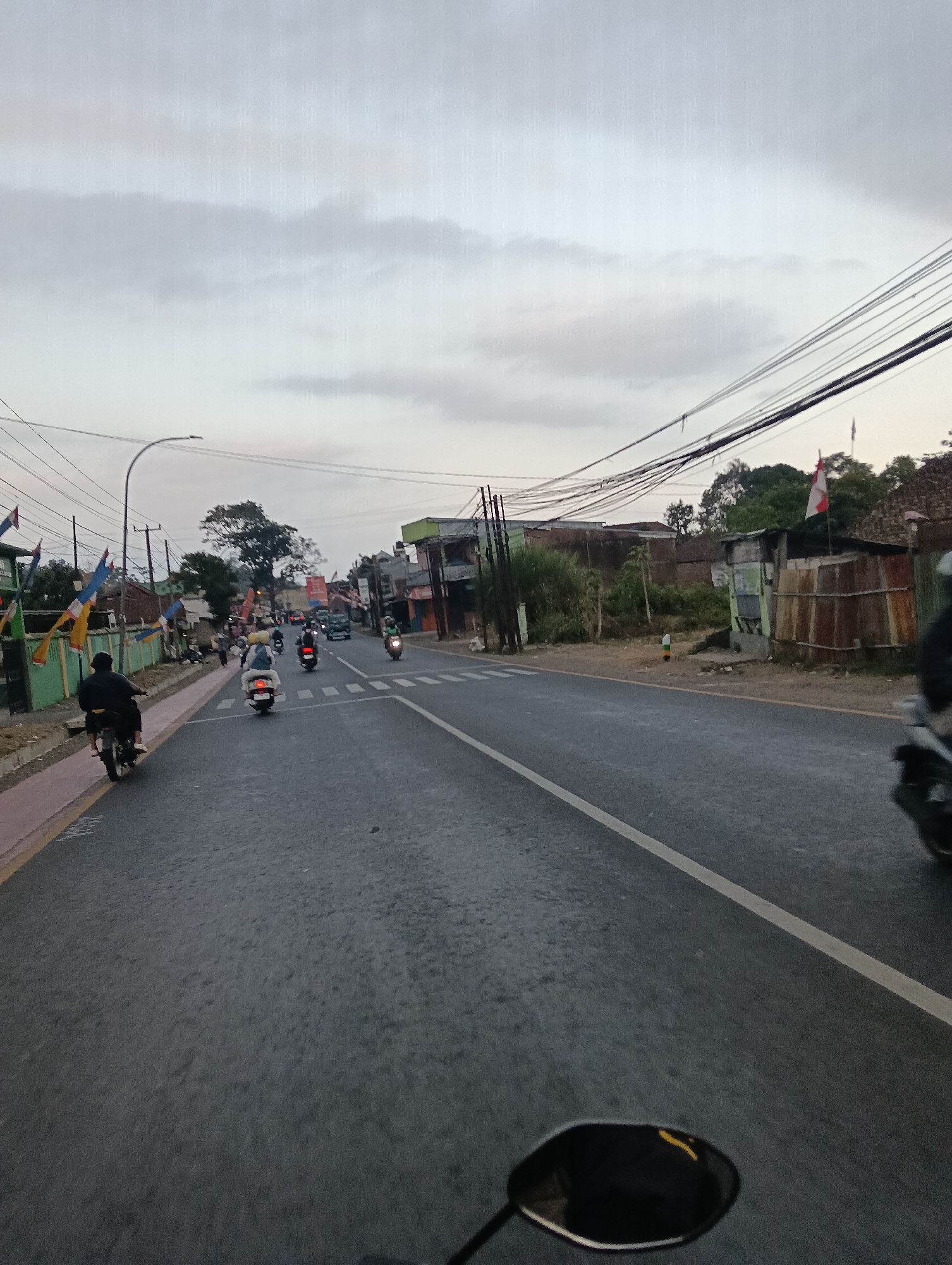 This image depicts a street scene, likely in Tasikmalaya, Indonesia, given the presence of Indonesian flags. The photograph is taken from the perspective of someone riding a motorcycle, as indicated by the motorcycle's rearview mirror visible in the foreground. The road is paved and appears to be moderately busy with several motorcycles and at least one car traveling in the same direction.
On the left side of the road, there is a pedestrian walkway with a red and yellow border. Buildings line the left side of the street, some with green facades and decorative flags. Beyond the buildings, trees and utility poles with numerous overhead wires are visible.
The right side of the road features a more rustic appearance, with a small, dilapidated structure made of corrugated metal and wood. An Indonesian flag is prominently displayed on a pole next to this structure. Further down the road, more buildings and trees can be seen.
The sky is overcast with gray clouds, suggesting it might be early morning, late afternoon, or a cloudy day. The overall lighting is subdued. The atmosphere appears to be that of a typical urban or semi-urban environment in Indonesia, characterized by motorcycle traffic and a mix of modern and older structures.