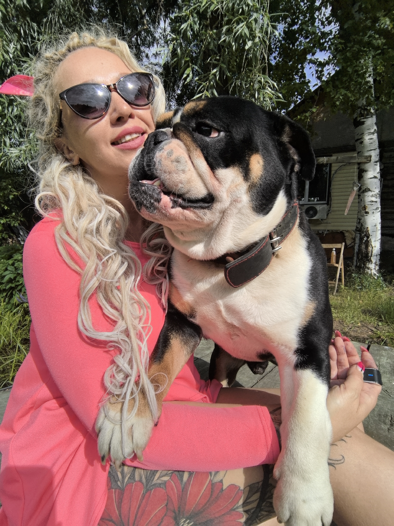 A woman with blonde curly hair and sunglasses is holding a tricolor bulldog. She is wearing a bright pink top and has tattoos on her legs. The dog is wearing a black collar with red trim. They are outdoors in a sunny environment, likely during the day. The background includes trees, foliage, and a building with a white birch tree in front of it. The overall mood appears relaxed and cheerful. There is no visible text in the image.