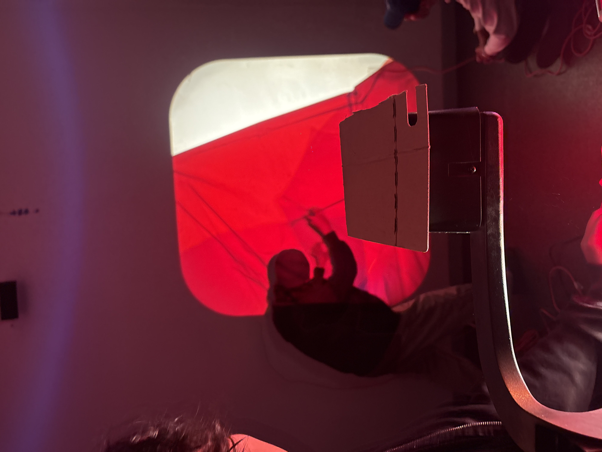 The image depicts an indoor scene in Mexico City, Mexico, illuminated by prominent red and purple ambient lighting. In the far-left foreground, the upper left portion of a person's head and shoulder are visible, showing dark, curly hair. Directly across the mid-ground, a dark, rectangular piece of equipment, likely a projector, stands with a light-colored, multi-tiered cardboard attachment on its top surface. This attachment features distinct notched cutouts.
On the back wall, a large, rounded rectangular projection displays a two-tone design: a red, wedge-shaped area on the left, and a white, curved area on the right. Against this projection, a standing person is silhouetted, appearing to manipulate a thin line or object with both hands at chest level. To the far right, a third individual is seated on the dark, reflective floor, wearing a blue cap and a light-colored long-sleeved shirt, actively handling or coiling a bundle of red cables or ropes. The floor reflects the red lighting, while the walls, originally light-colored, are tinted with the red and purple hues of the illumination. The scene conveys a technical or artistic setup involving projections and equipment management.