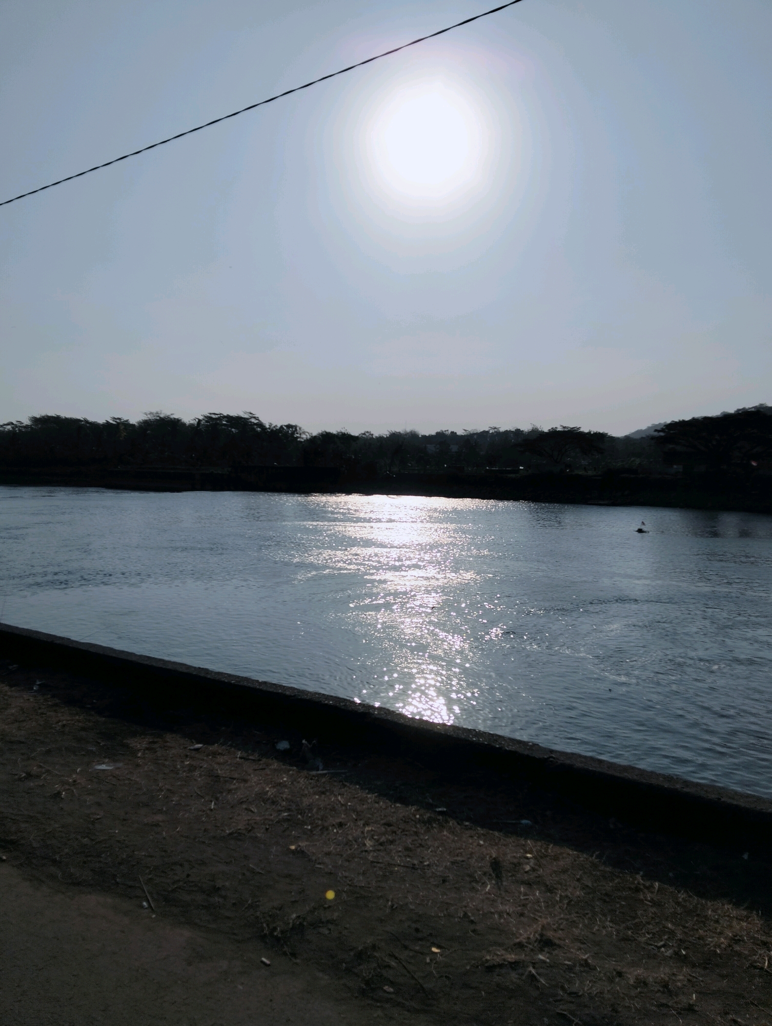 The image depicts a wide river flowing through a natural landscape. In the foreground, a dark, textured embankment lines the river. The river's surface is choppy, reflecting the bright, hazy sun that dominates the upper portion of the image. A thin black wire stretches diagonally across the sky. Silhouetted trees form a dense line along the opposite bank of the river, suggesting a forested area. In the distance, hills are faintly visible. A small, dark object, possibly a bird or a buoy, is present in the water towards the right side of the frame. The overall scene is characterized by natural elements under a bright, overcast sky. The location is specified as the city of Kebasen, Indonesia.