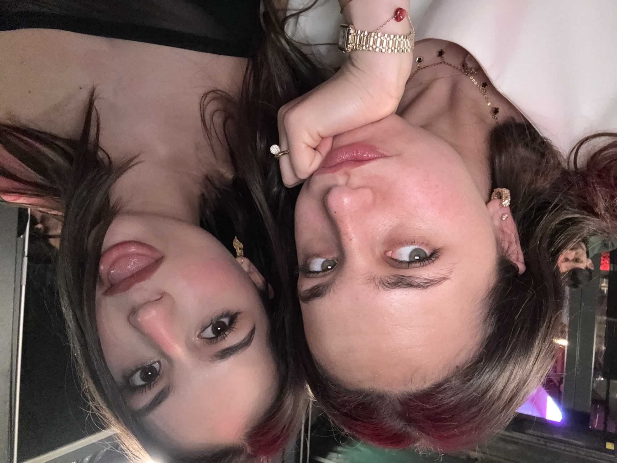 Two fair-skinned women pose for a selfie. The woman on the left has dark brown hair with red highlights, her tongue is slightly sticking out of her mouth, and she wears dark eyeliner and gold dangling earrings. The woman on the right has dark brown hair with red highlights and wears a gold watch, ring, bracelet, gold earrings, and a gold necklace with black stars. She is resting her face in her hand. The background appears to be a bar or restaurant, with a man wearing a mask and several windows visible. There is no definitive information to determine that this image was taken in Milan, Italy.