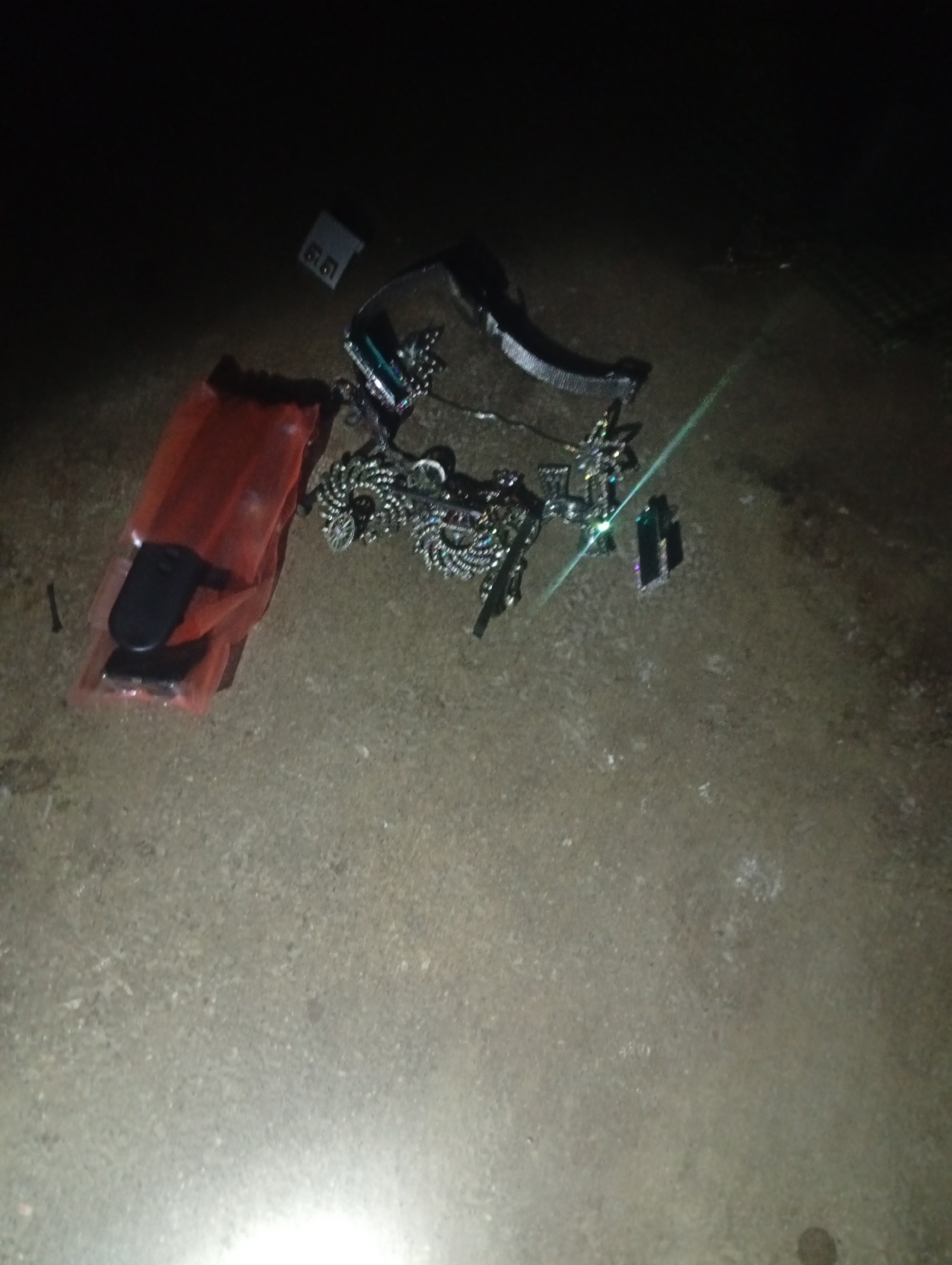 A close-up, low-light, outdoor shot captures various objects scattered on a rough, concrete surface, possibly in Jalingo, Nigeria. In the foreground left, a red plastic bag contains two black electronic devices, possibly key fobs. To the right of the bag, a tangled assortment of shiny, metallic chains and what appear to be decorative charms or pendants are spread across the ground. Further to the right, a small, dark rectangular object is visible. The background is predominantly dark, with some faint light reflections suggesting artificial lighting. The surface appears to be a dusty, uneven concrete pavement.