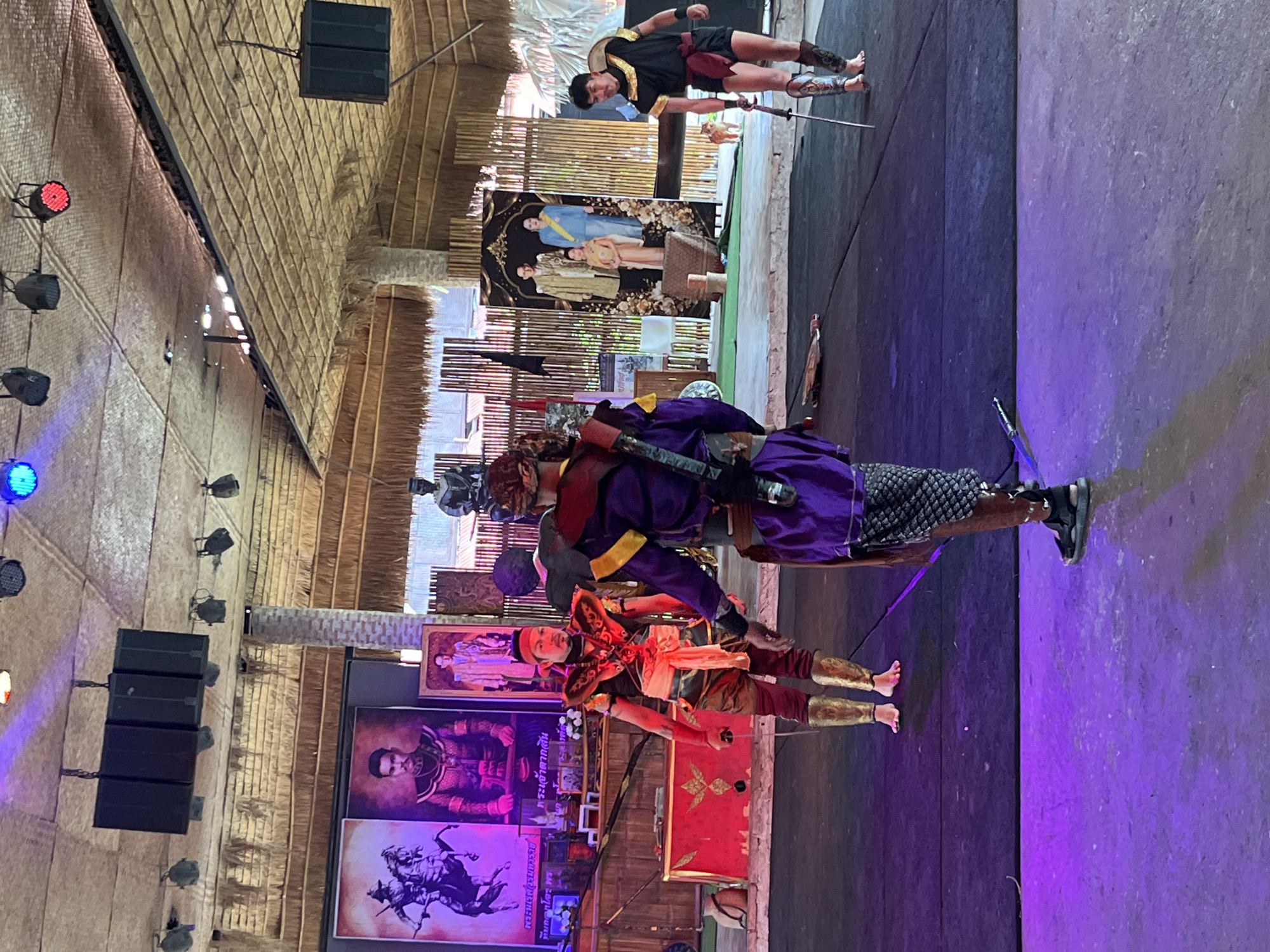 This image captures a scene of a traditional Thai performance taking place in Ayutthaya Town Municipality, Thailand. Three men, dressed in ornate warrior costumes, are present on a dark performance stage. The man in the foreground is wearing a purple tunic, patterned shorts, and ornate leg guards. He is facing away from the camera, holding a sword. To his left, another performer in a red and gold costume with gold leg guards is holding a sword and looking towards the center. A third performer, wearing a black and gold costume with red trim and leg guards, stands to the far right, also holding a sword.

Behind the performers, a backdrop displays historical portraits. On the left, a large portrait depicts a man on horseback, and next to it, a portrait of a man in regal attire. A smaller portrait of a figure in a gold robe is also visible. Further back, another backdrop features a formal portrait of a royal family. The performance space is covered by a thatched roof, supported by bamboo structures, and illuminated by stage lights casting purple and blue hues. Speakers are mounted on the ceiling. The overall setting suggests an outdoor or semi-outdoor venue designed for cultural presentations.