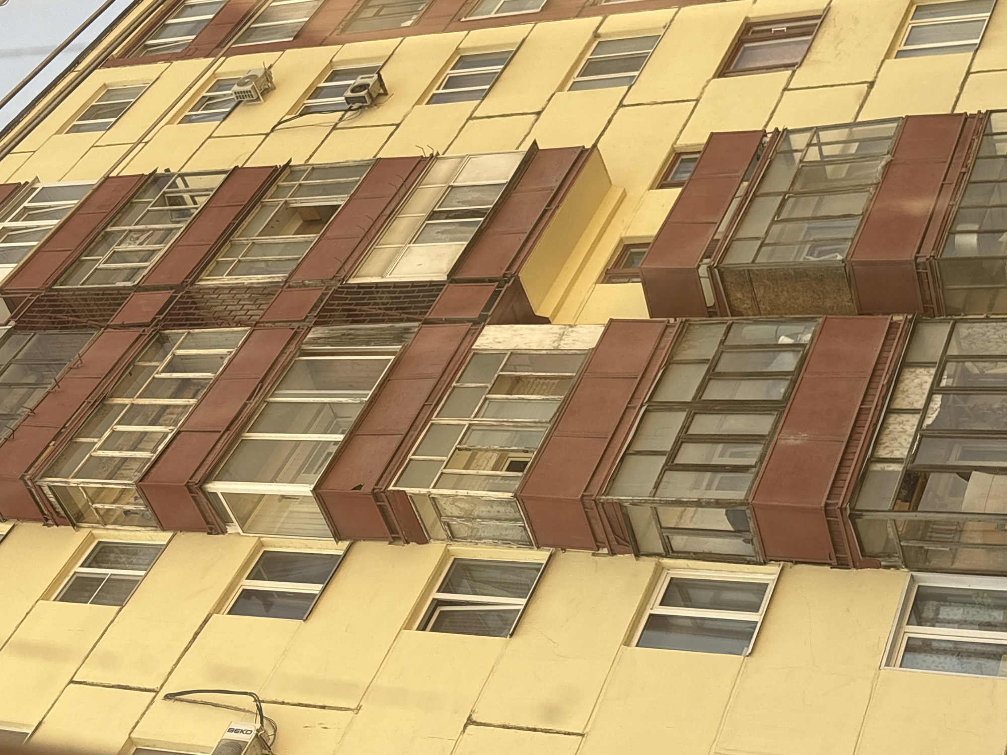 The image depicts a section of a building in Izhevsk, Russia. The exterior wall has a yellow tone. The facade is composed of a repetitive pattern of windows. Some windows have projecting, box-like structures, typically colored in reddish-brown. A few air conditioning units are mounted on the building's surface. Some of the windows appear damaged. A "BEKO" branded air conditioning unit is visible in the lower section of the image.