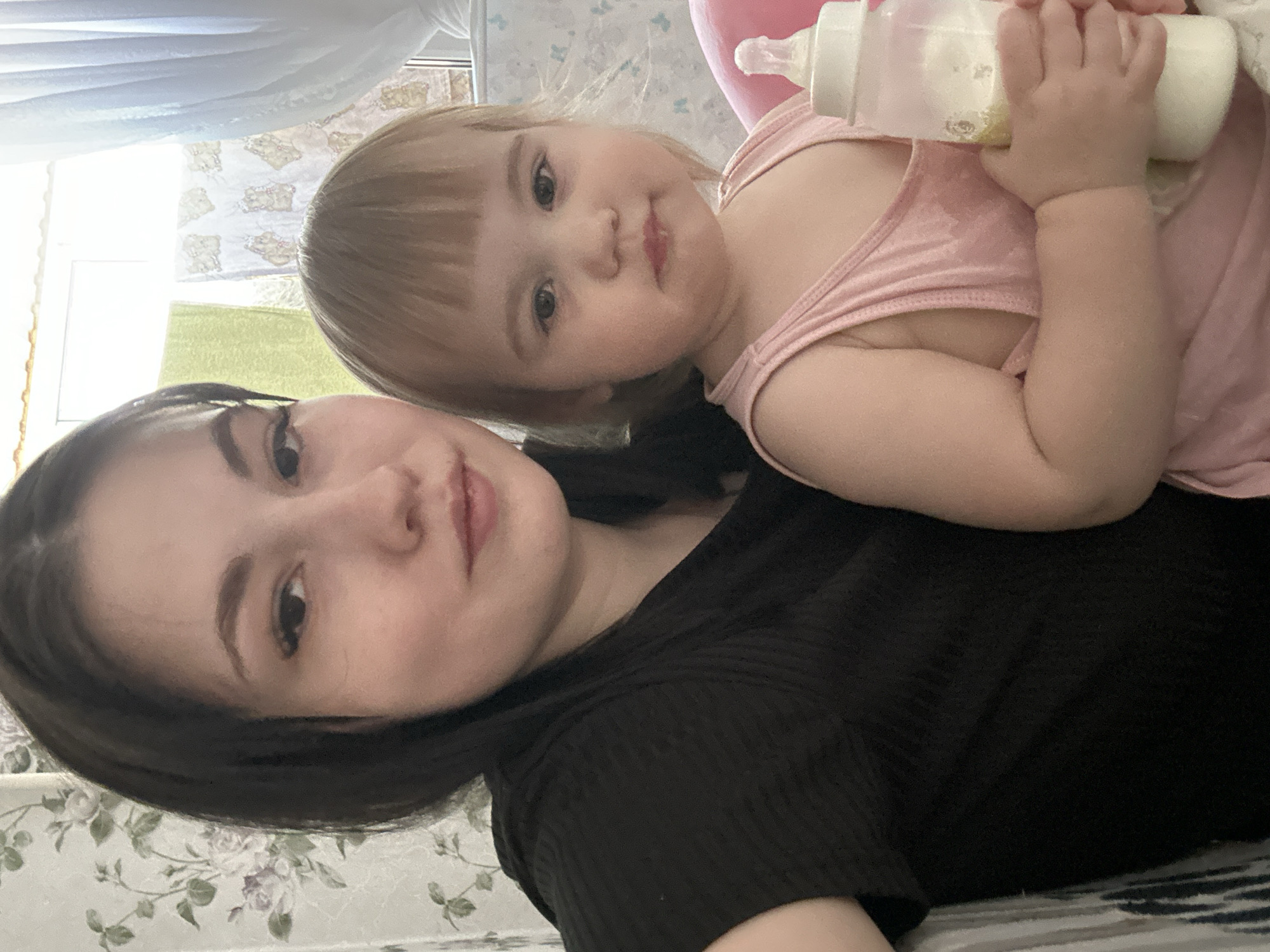 A selfie captures a woman with dark hair and a baby holding a bottle. The woman is wearing a black shirt, and the baby is dressed in pink. The background shows a patterned wallpaper and a glimpse of curtains. The photo was likely taken indoors. The location is Chaikovsky, Russia.