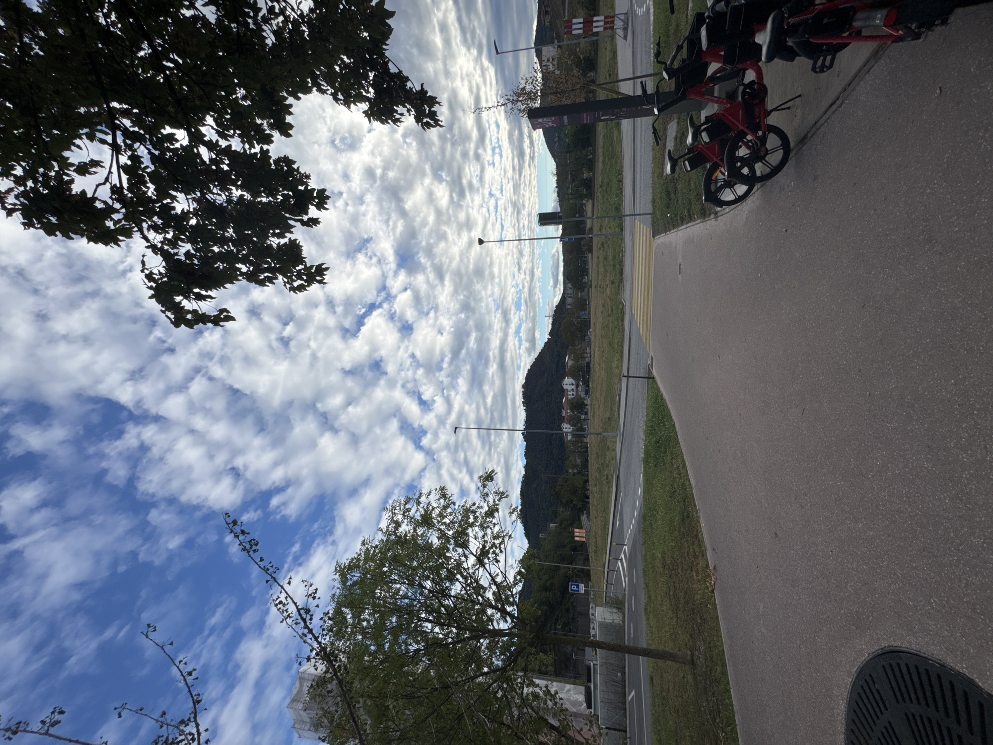 The image shows a paved walkway in the foreground, with several red shared bicycles parked on a concrete curb to the right. Beyond the walkway is a road with a pedestrian crossing, and a grassy area. In the background, there are trees, buildings, streetlights, and a large hill under a sky with scattered clouds. The location is identified as Morbio Inferiore, Switzerland.