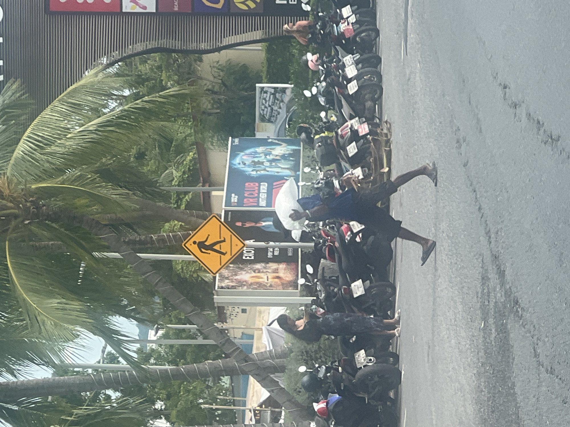 A pedestrian crosses a paved area in front of a line of parked motorbikes. The pedestrian, wearing a blue shirt and dark shorts, is carrying a white object over their head. To the left, a woman in a dark patterned dress stands looking at a mobile phone. Behind the motorbikes are billboards, including one for "VR CLUB ANOTHER WORLD." Palm trees with visible wrapped trunks are present in the background. A yellow pedestrian crossing sign is visible above and to the left of the woman. The environment appears to be an outdoor area, likely a parking lot or thoroughfare, in a tropical or subtropical climate, possibly located in Ban Bang Rak, Thailand.