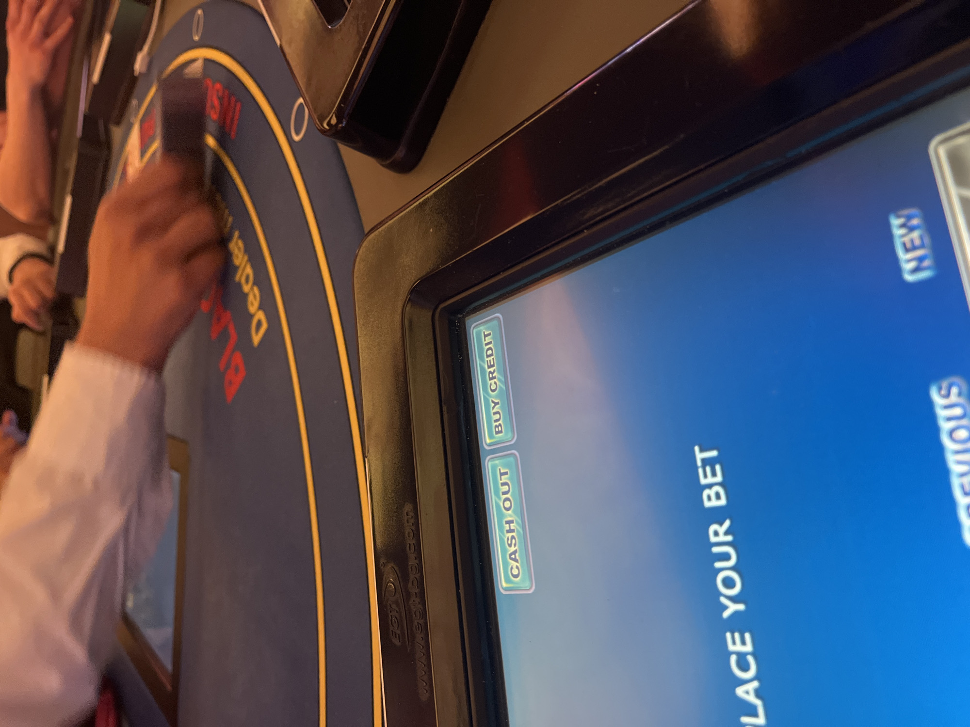The image shows a close-up of an electronic blackjack table in what appears to be a casino. A person wearing a white shirt is visible to the left, with their hand near the table surface. The table has a screen display showing "PLACE YOUR BET" along with buttons for "CASH OUT," "BUY CREDIT," "NEW," and "PREVIOUS." The rim of the table, labeled "Blackjack Dealer..." is also visible. There is no indication of the exact location in Burgas, Bulgaria.