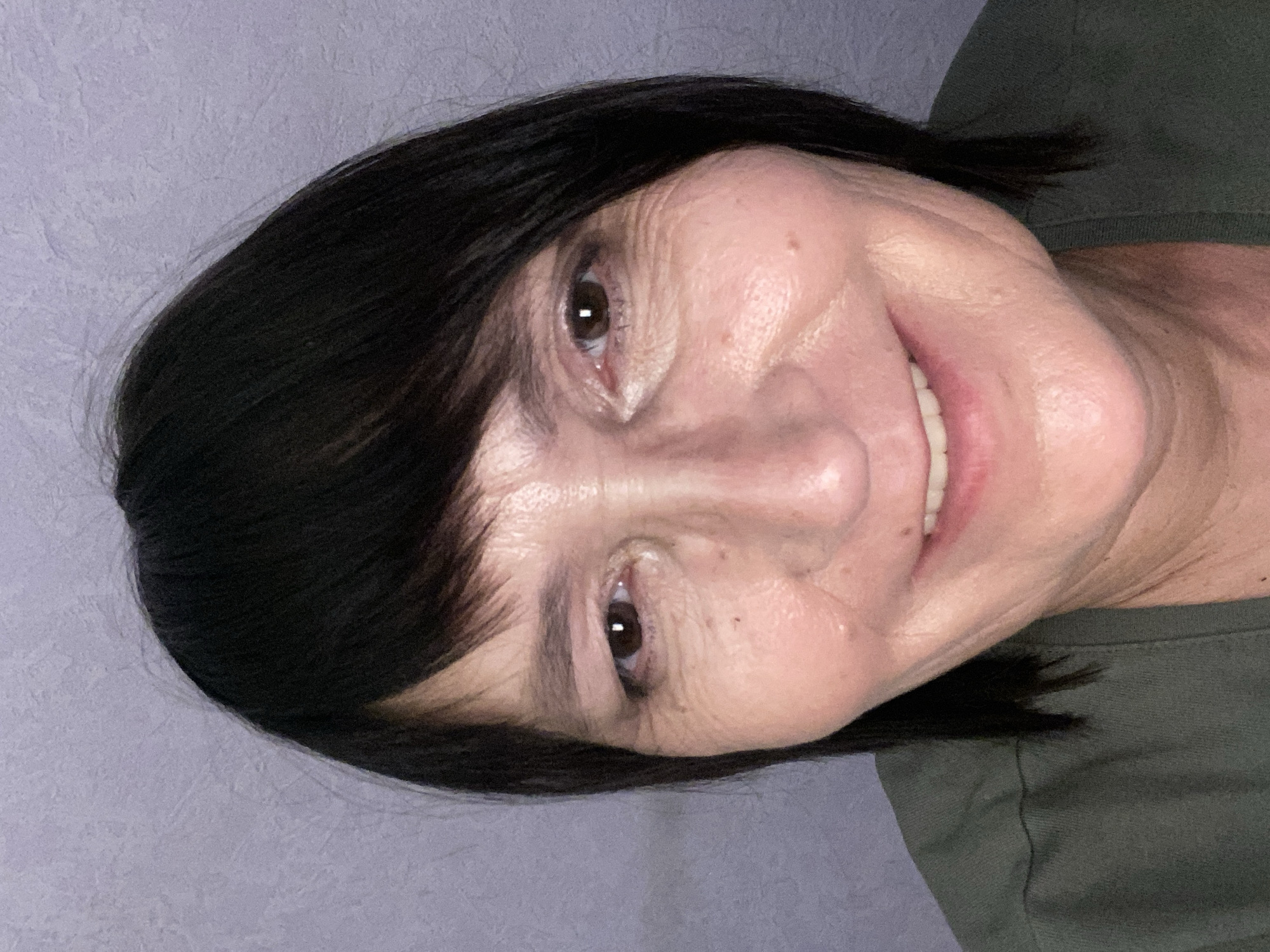 This media file features a close-up, head-and-shoulders shot of a woman with dark hair and fair skin, with her head tilted. She is wearing a dark green t-shirt. Her eyes are open and looking towards the viewer, and she has a smile on her face. The background is a textured, light purple wall. The image appears to be a selfie, likely taken indoors, with soft lighting. There is no visible text in the image. The location context provided is Novokuznetsk, Russia.