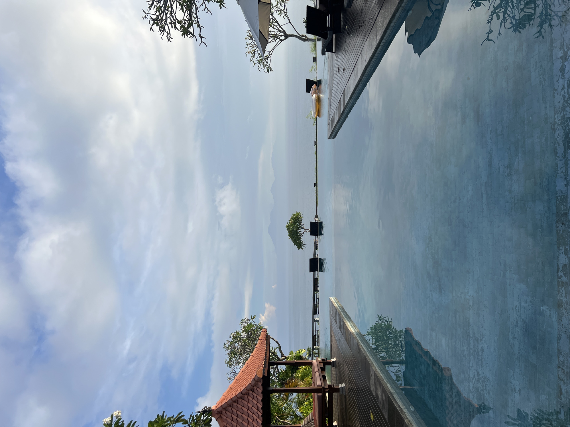 This image captures a serene and luxurious resort setting in Kuta Selatan, Indonesia, featuring an infinity pool overlooking a vast body of water and distant mountains under a partly cloudy sky.
In the foreground, an infinity pool with calm, reflective water dominates the lower half of the frame. To the right, a wooden deck extends alongside the pool, furnished with two dark lounge chairs. One chair is partially shaded by a white umbrella. A person is seen relaxing on a golden/yellow inflatable ring float in the pool, near the edge of the wooden deck. On the left side of the foreground, a building with a distinctive terracotta-tiled roof and wooden structural elements is partially visible, surrounded by lush green foliage. More green tree branches and leaves are visible in the upper corners of the frame.
Beyond the infinity pool, a wide expanse of still water, likely a large lake, stretches towards the horizon. A small, neatly trimmed tree stands on a platform or small island within this body of water, centrally positioned in the mid-ground. In the far distance, faint outlines of multiple mountain ranges are visible, blending softly into the hazy sky.
The sky above is bright, covered with scattered white and light gray clouds, suggesting it is daytime, possibly late morning or early afternoon. The overall impression is one of tranquility, leisure, and natural beauty. There is no visible text in the image.