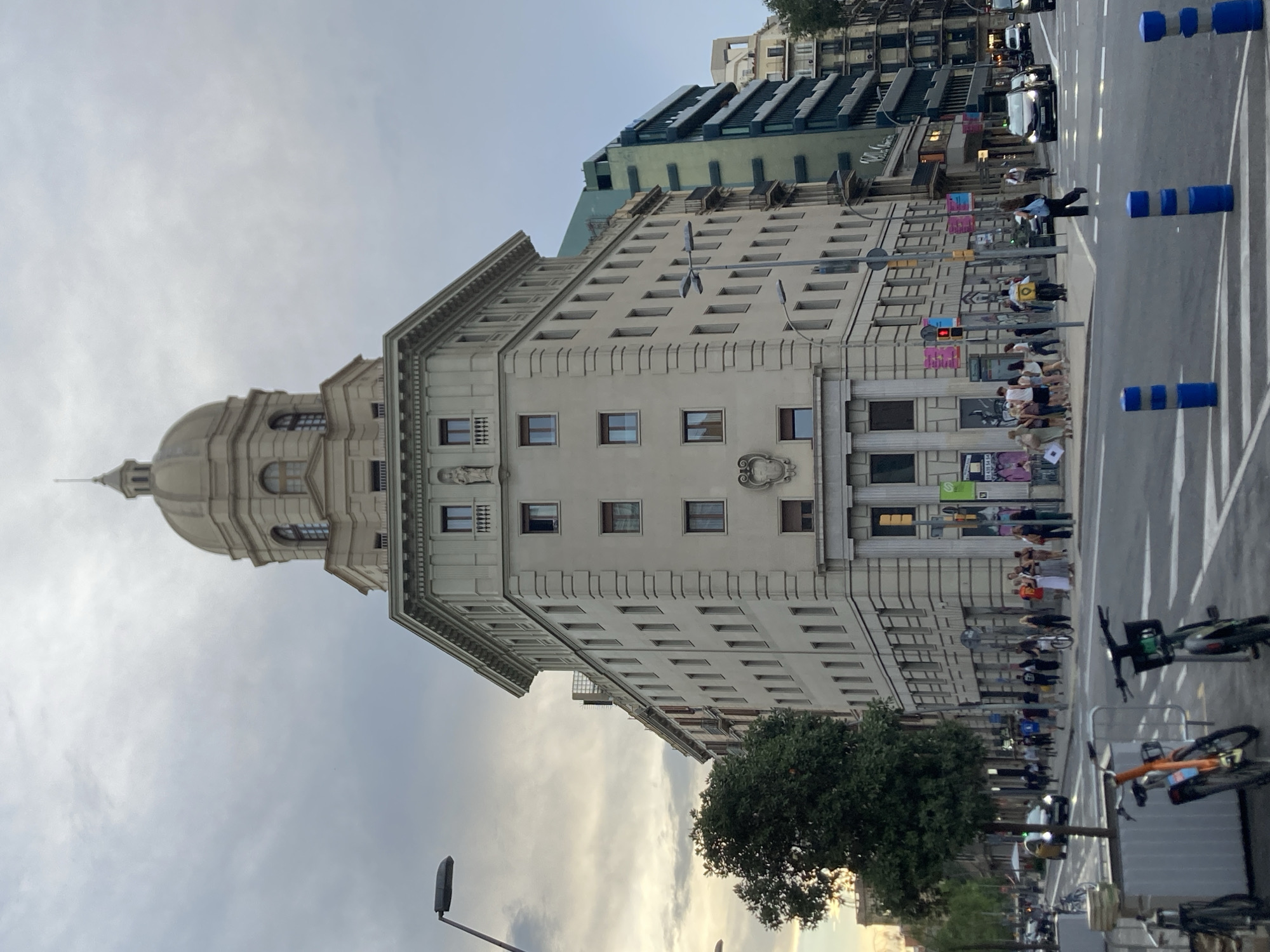 The image depicts a view of the building at Plaça de Catalunya in Barcelona, Spain. The building is characterized by a prominent dome on its top, classical architectural elements, and multiple floors with numerous windows. In the foreground, a street intersection is visible with pedestrian crossings, traffic lights, and streetlights. Several individuals are gathered on the sidewalk, some appearing to be tourists or locals. Traffic, including cars and a delivery person on a scooter, is present on the street. The sky is overcast with clouds, suggesting it is either early morning or late afternoon. To the right of the main building, other urban structures and buildings are visible, contributing to the dense city environment. A tree stands to the left, partially obscuring the lower portion of the main building. Several bicycles are parked along the sidewalk in the immediate foreground.