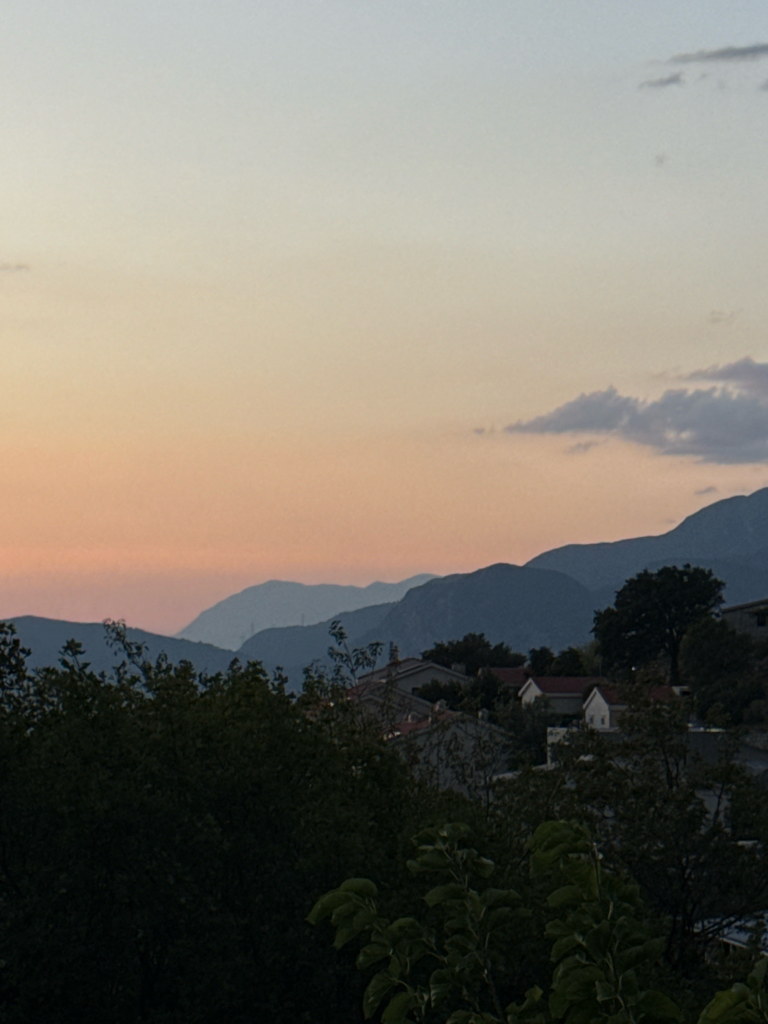 The image depicts a twilight sky with a gradient of soft orange and pale blue, transitioning into a darker blue at the horizon. Silhouetted against the sky are multiple layers of mountains. In the foreground, dense green foliage partially obscures a cluster of buildings with red-tiled roofs, characteristic of the town of Rađenovići, Montenegro. The overall impression is a peaceful, serene landscape at dusk.