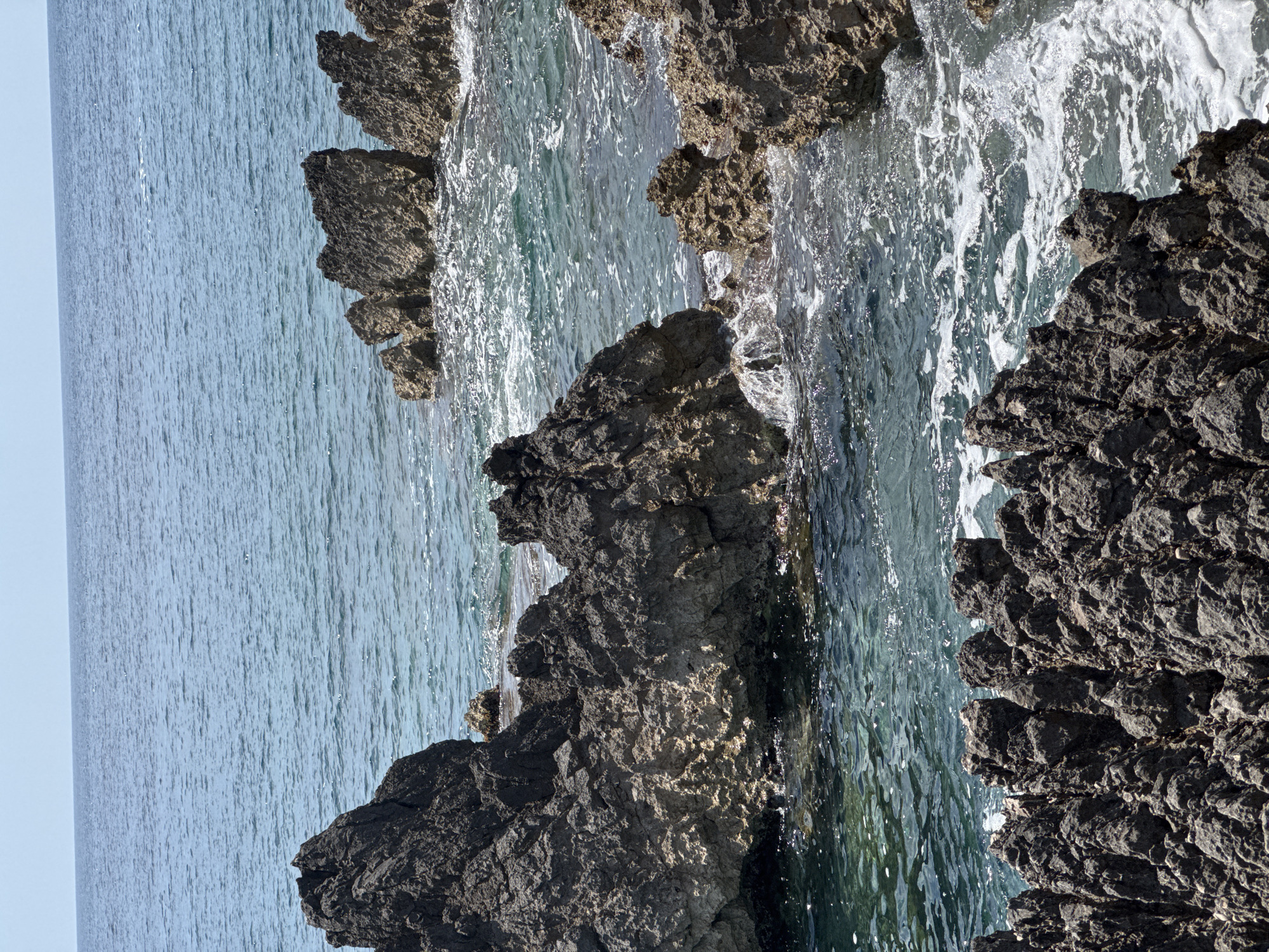 The image depicts a rocky coastline in Dubraljevina, Montenegro, where dark, jagged rocks meet the turquoise-colored sea. The water shows signs of waves crashing against the rocks, producing white foam and splashes. The rocks are of varying sizes, some large and dominating the foreground, others smaller and receding into the distance. The sky is a pale blue, suggesting a clear day. The composition is such that the rocks are prominently featured, with the sea serving as a backdrop, creating a natural coastal landscape.