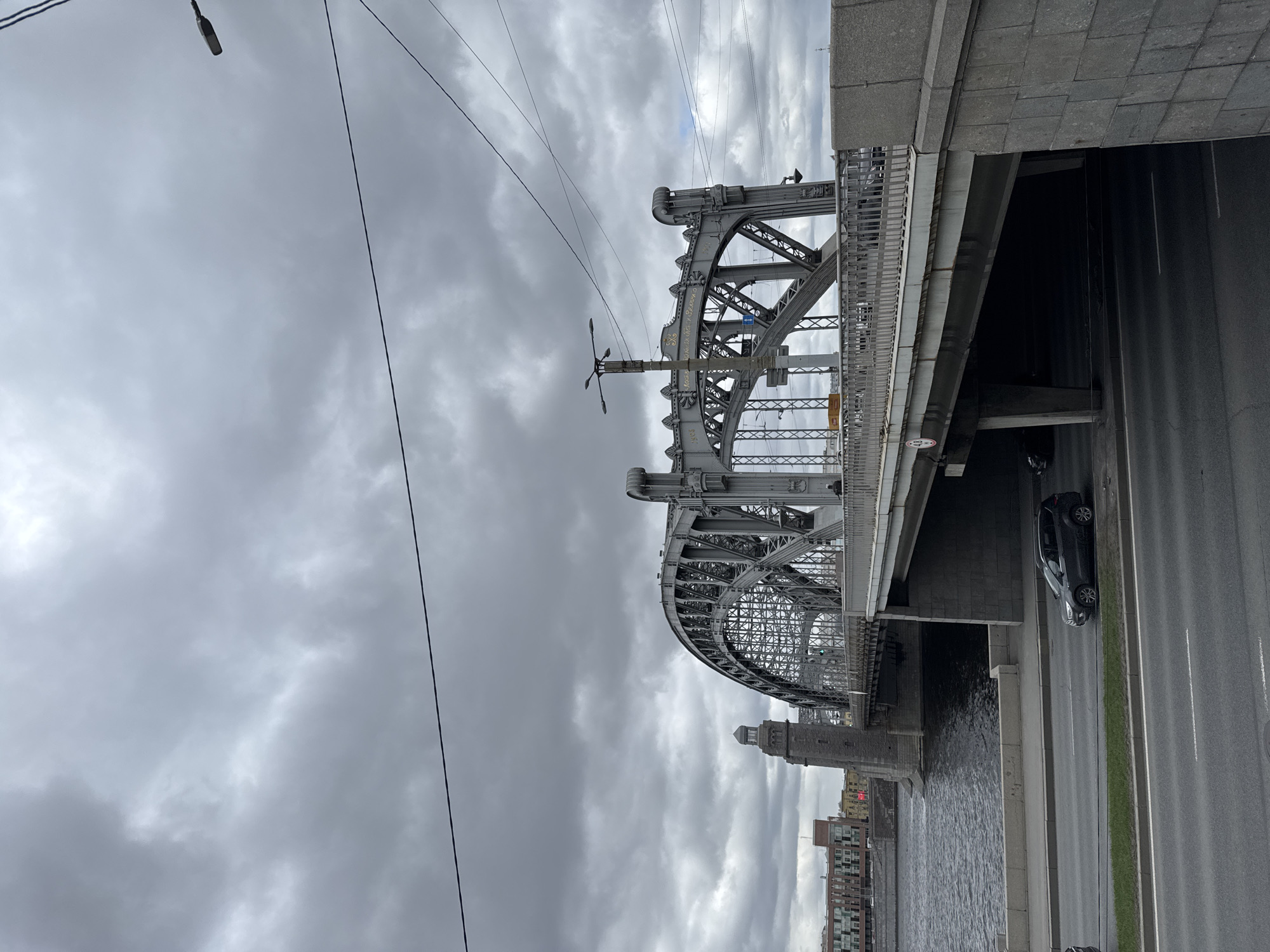 The image shows the Peter the Great Bridge, a drawbridge across the Neva River in Saint Petersburg, Russia, under a cloudy sky. The bridge is constructed of steel with an arched design. A road runs alongside the river, with at least one car visible. Buildings are visible in the background on the opposite side of the river. A street lamp and overhead wires are visible in the upper left portion of the image. The river is calm with a slight ripple on the surface.