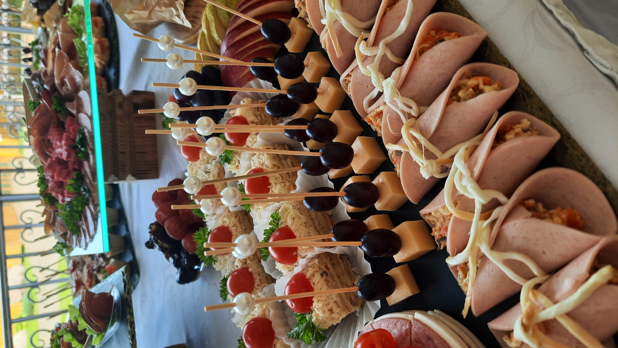 An overhead medium shot captures a festive spread of appetizers on a table, set outdoors in what appears to be a rural setting. The cuisine is varied, featuring an assortment of cold cuts, sliced meats, and cheeses arranged on platters. Bite-sized snacks are prominently displayed, with small sandwiches topped with cherry tomatoes and parsley, skewers of grapes and cheese cubes, and meat rolls filled with a creamy mixture and tied with cheese strings. The background hints at a scenic view with a wrought-iron railing and greenery, suggesting an event held in a picturesque location in the city of Жодзішкаўскі сельскі Савет, Belarus. The lighting indicates it's daytime, possibly late morning or early afternoon, given the bright, natural light illuminating the scene. The focus is on the food, presented in an appetizing and organized manner, suitable for a gathering or celebration.