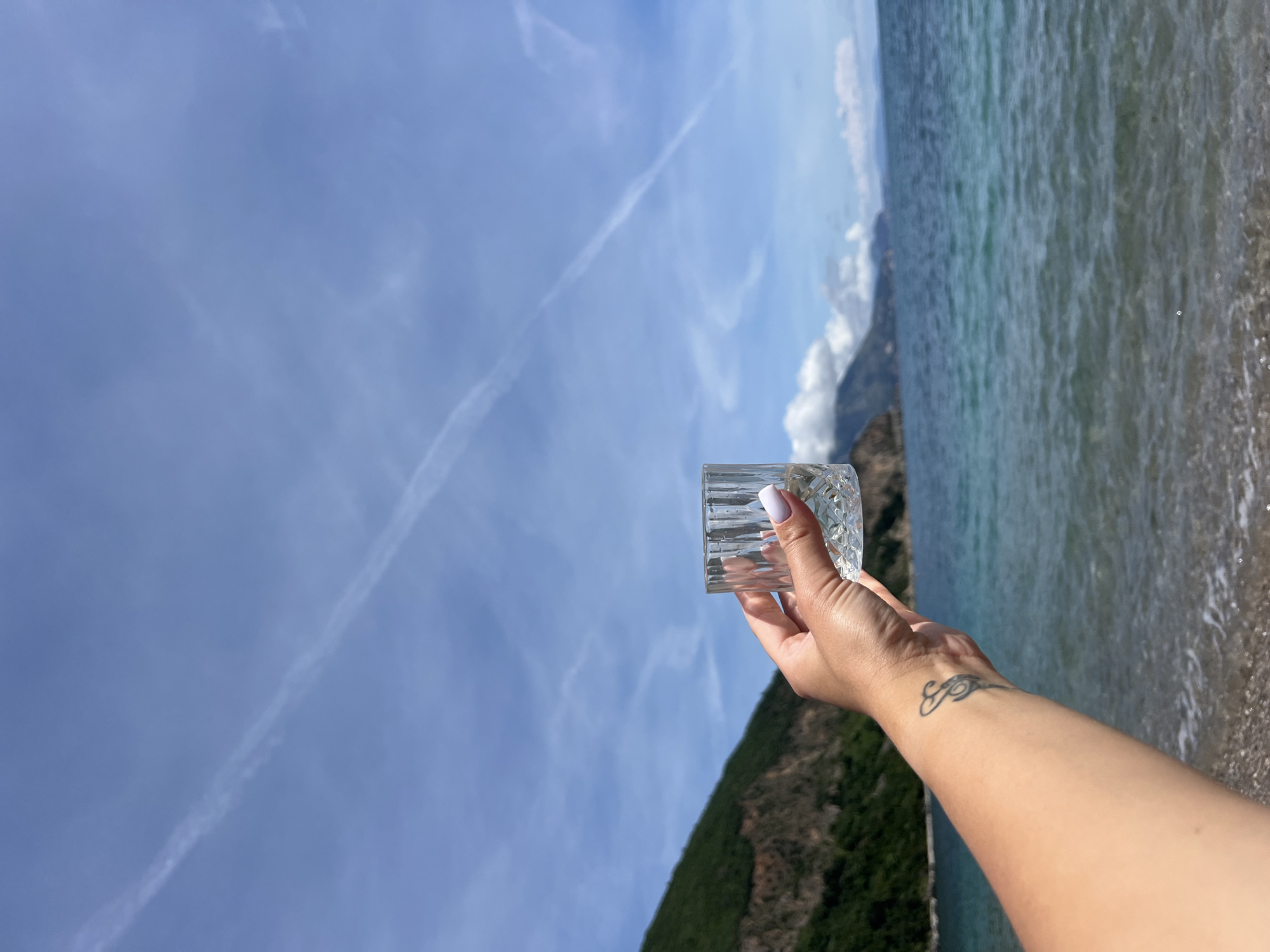 The image depicts a woman's hand holding an empty, ornate glass in an outdoor setting. The hand, with white nail polish and a tattoo on the wrist, is positioned against a backdrop of a blue sky with contrails, a turquoise sea, and a green, mountainous shoreline. The location is likely near Prijevor, Montenegro, given the coastal and mountainous landscape. The focus is on the glass and the hand holding it, suggesting a moment of contemplation or a toast. The environment consists of a clear sky, a scenic coastline, and lush greenery.
