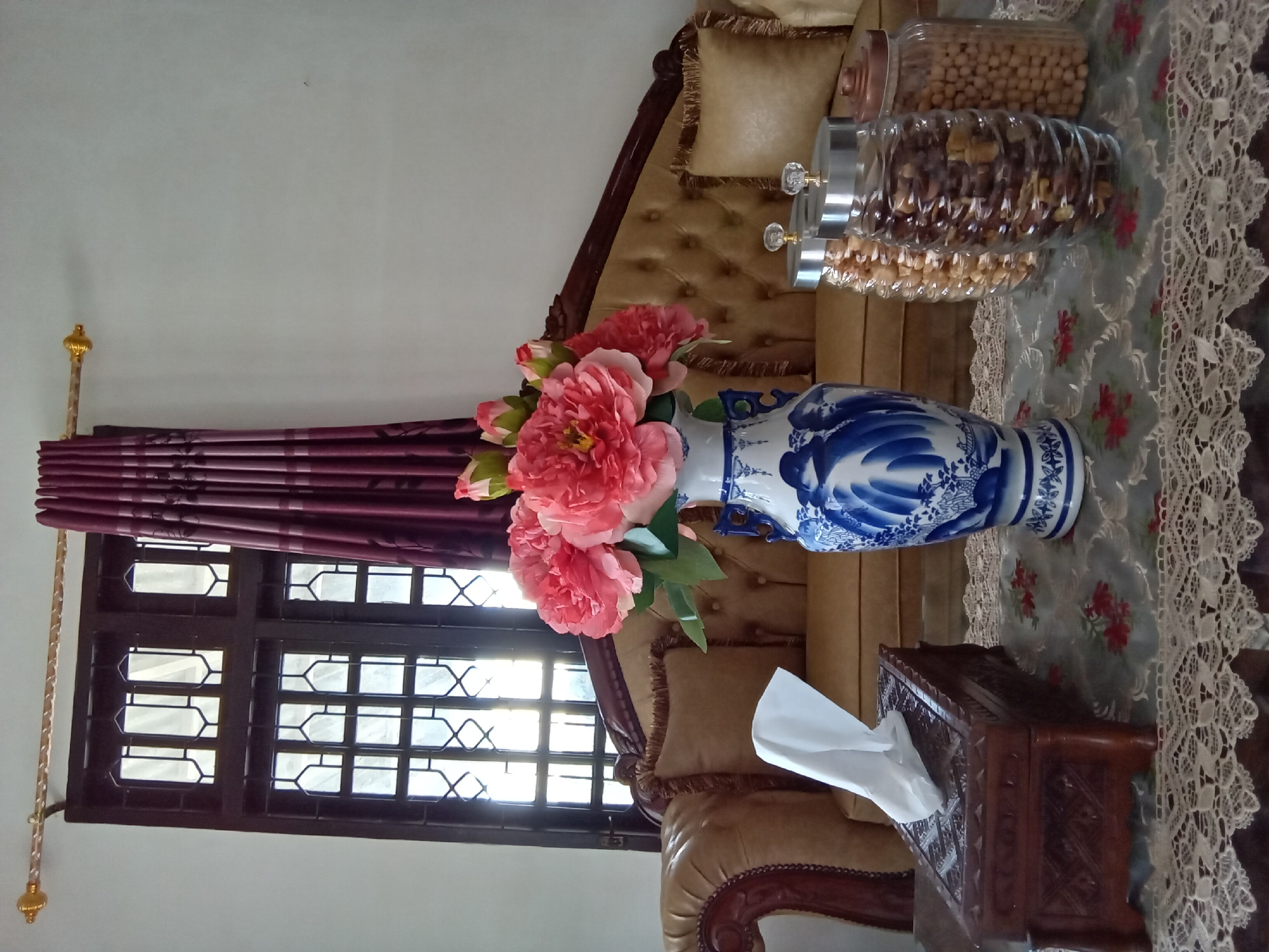 This interior photograph, taken in Lhokseumawe, Indonesia, depicts a segment of a living space. The scene is dominated by a traditionally styled, light brown tufted sofa with dark wooden accents and matching fringed throw pillows, positioned against a plain, light-colored wall. A large, dark brown wooden window frame with intricate metal security bars allows bright daylight to enter from the left, illuminating the room. A dark red/maroon curtain with a subtle pattern is drawn to the right side of the window, hanging from a golden, ornate curtain rod.
In the foreground, a dark wooden table is covered by a white lace tablecloth, overlaid with a blue-gray fabric featuring red floral patterns. Centrally located on the table is a tall, blue and white ceramic vase, decorated with traditional landscape or figure motifs, holding a bouquet of artificial pink peony flowers and green foliage, including several closed buds. To the left of the vase, a dark brown, carved wooden tissue box holder with a single white tissue protruding is visible. To the right of the vase, three glass jars containing various snacks are arranged. The leftmost jar, partially visible, contains light-colored snacks; the middle, fluted jar contains darker brown snacks; and the rightmost cylindrical jar contains light brown, spherical snacks, likely chickpeas. The first two jars have silver lids with clear, decorative knobs, while the third has a copper-toned lid. No individuals are present in the image.