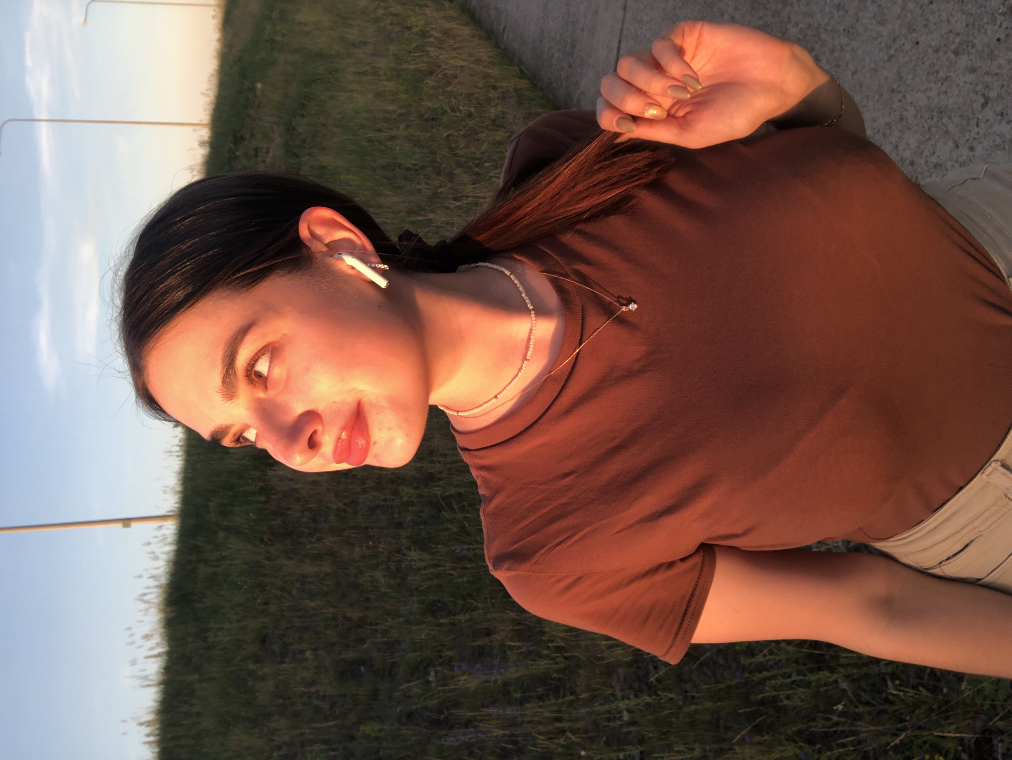 A young woman with dark hair pulled back in a ponytail is captured in a close-up, outdoor shot. She is wearing a brown t-shirt and light-colored pants. Her left arm is bent and resting behind her head, with her hand visible, showcasing manicured nails with a subtle shimmer. She wears a delicate necklace with a small pendant and white earbuds. Her head is tilted, and she looks off to the left, her expression seemingly contemplative. The lighting suggests it is either sunrise or sunset, casting a warm, golden glow on her face and skin. The background features a grassy area and a paved surface, possibly a sidewalk or path, with streetlights visible in the distance. The sky is a pale blue, indicating clear weather. The scene evokes a sense of calm and introspection.