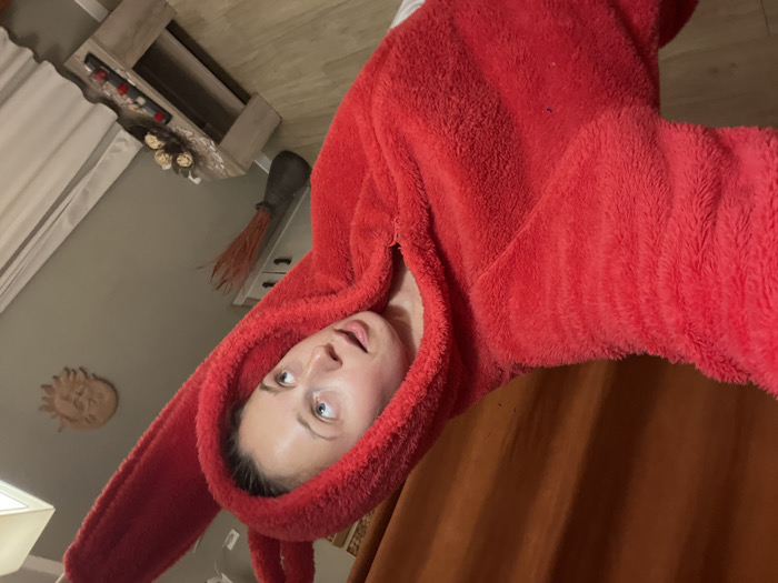 A young adult female is depicted in a selfie, taken indoors in Boreti, Montenegro. She has fair skin, light-colored eyes, and her hair is pulled back. She wears a bright red, fuzzy hooded jumpsuit, which features prominent rabbit-like ears on the hood. Her mouth is slightly open, and her gaze is directed towards the viewer's upper left. Her right arm, holding the camera, is extended towards the viewer. The background features a neutral gray wall, with dark orange curtains to the left and light-colored curtains to the right. A white cabinet with dark handles is visible behind her left shoulder. On the gray wall, a light-colored, circular sun-face decorative piece is mounted. Below it, a light-toned, low-profile coffee table holds a tray with several spherical decorative items and a rectangular object with red accents. A dark vase with dried reddish plant material is on the floor next to the cabinet. The floor is light-colored wood or laminate, and a rectangular ceiling light fixture is visible at the top right.