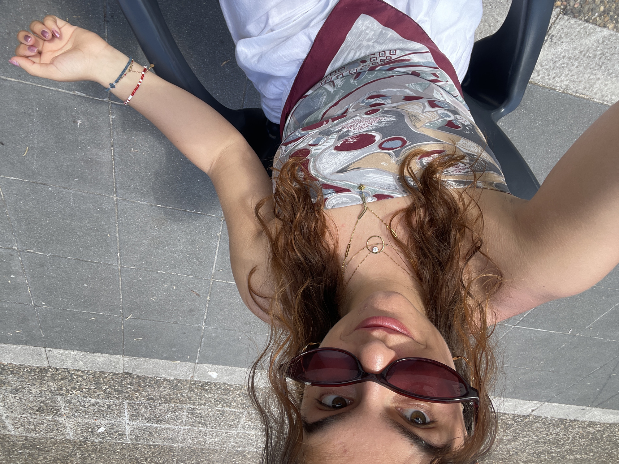 The image shows a woman sitting on a black plastic chair in Jerez De La Frontera, Spain. She is wearing white clothing and a patterned top with maroon trim. The ground is paved with gray tiles. The woman is facing the camera, wearing sunglasses, and has necklaces visible. She is wearing bracelets on her left wrist.