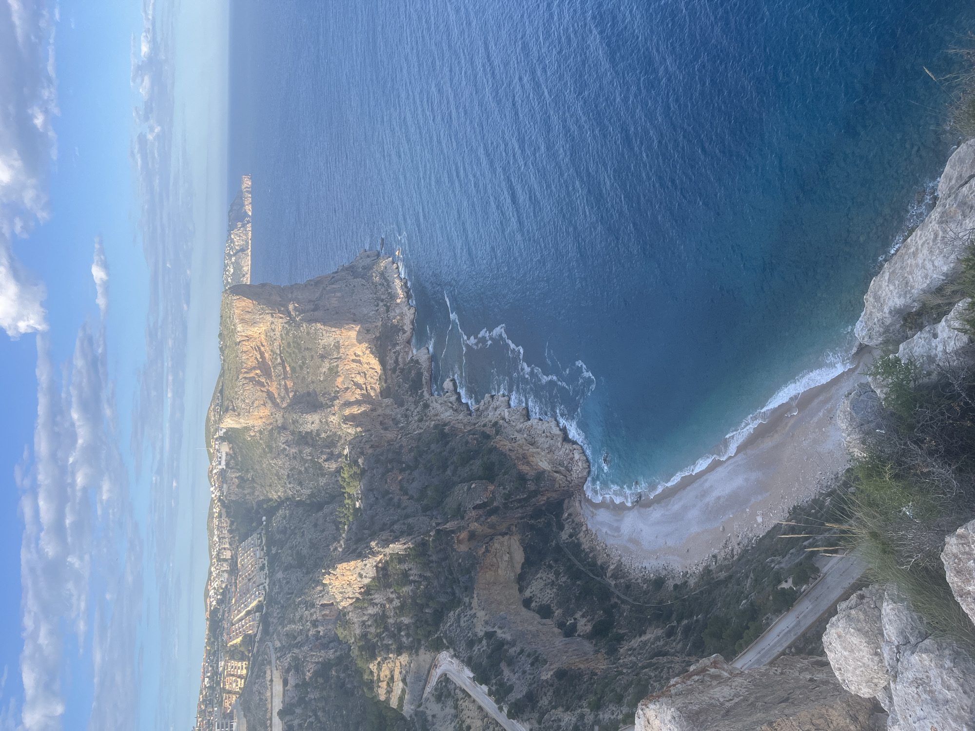 The image captures a high-angle view of the coastline in el Poble Nou de Benitatxell/Benitachell, Spain. The scene encompasses the cliffs, a beach, the ocean, and part of the town.
Cliffs: A large cliff dominates the frame, displaying rocky textures and sparse vegetation.
Beach: A beach with light-colored sand stretches along the base of the cliff. The shoreline is defined by waves meeting the sand.
Ocean: The deep blue ocean extends to the horizon.
Town: A portion of the town with residential buildings and a road winding along the cliffside are visible.
Sky: The sky is blue with scattered clouds.