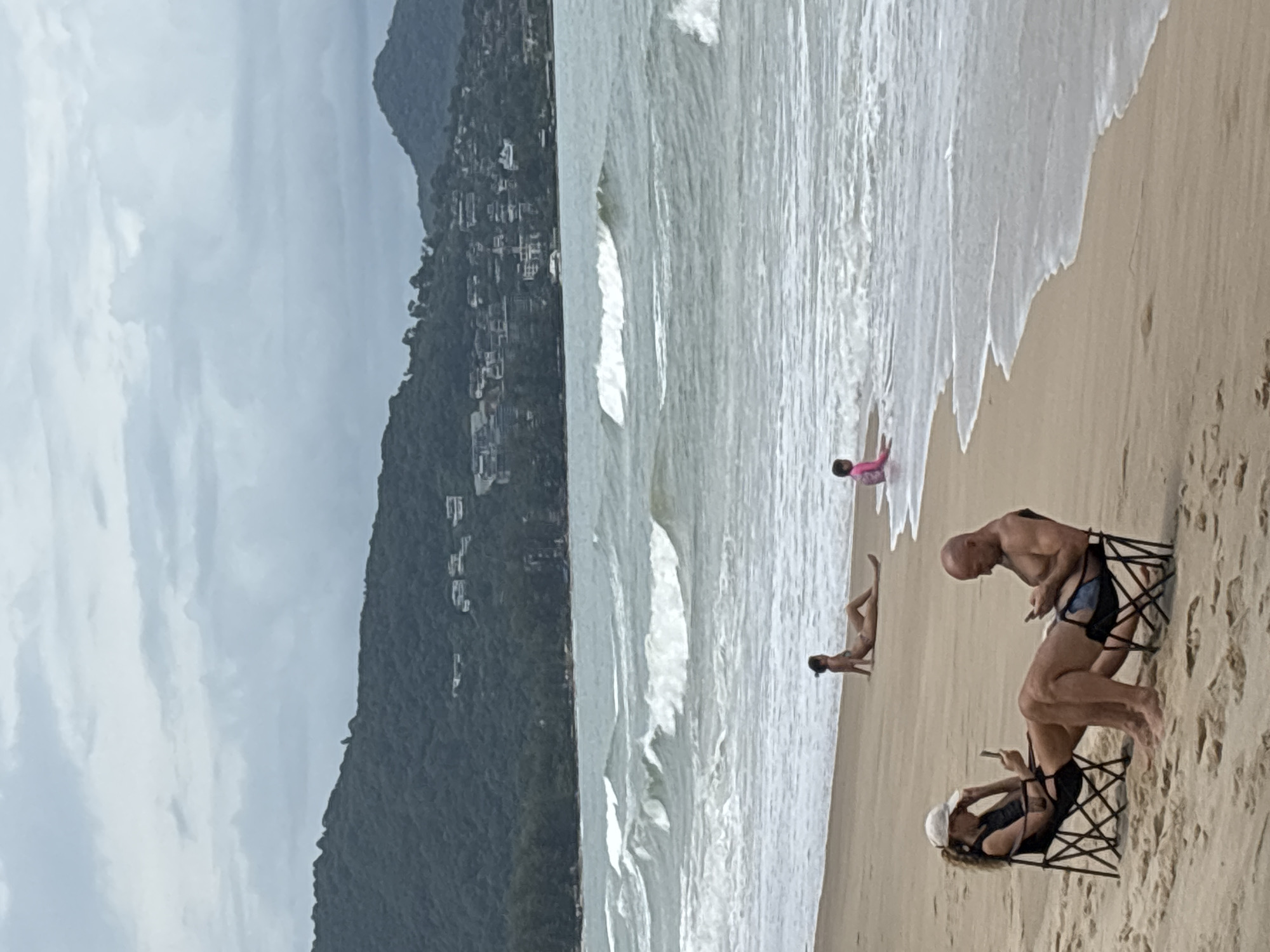 The image depicts a beach scene in Choeng Thale, Thailand, on an overcast day. The water is rough, with waves breaking near the shore. A child in a pink shirt is standing in the surf. Further down the beach, a person is lying on the sand. Two people sit in beach chairs on the sand, looking at their phones. Behind the beach is a mountain with buildings.