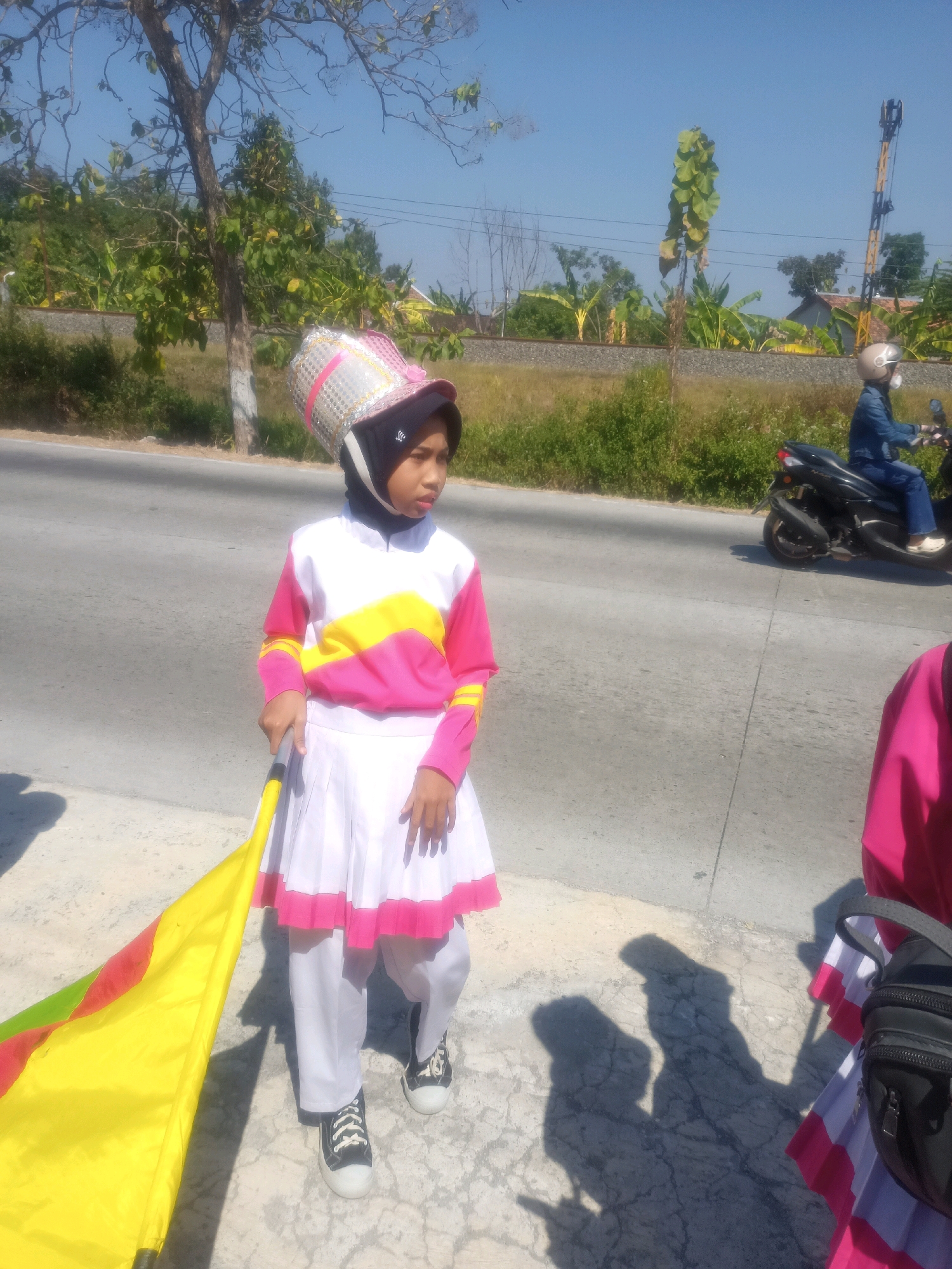 In Sumberlawang, Indonesia, a young girl in a colorful marching band uniform stands on a paved road under a clear blue sky. Her uniform consists of a pink, white, and yellow long-sleeved shirt, a white pleated skirt with a pink trim, and white pants. She wears a black hijab and a decorative, tall hat adorned with silver sequins and pink accents. She holds a flag with yellow, red, and green sections. A person on a black motorcycle, wearing a blue denim jacket and a helmet, rides on the road in the background. The environment includes a tree with bare branches and green leaves, a railway track with a signal pole, and vegetation on the side of the road. The sun casts shadows of people and objects on the ground.