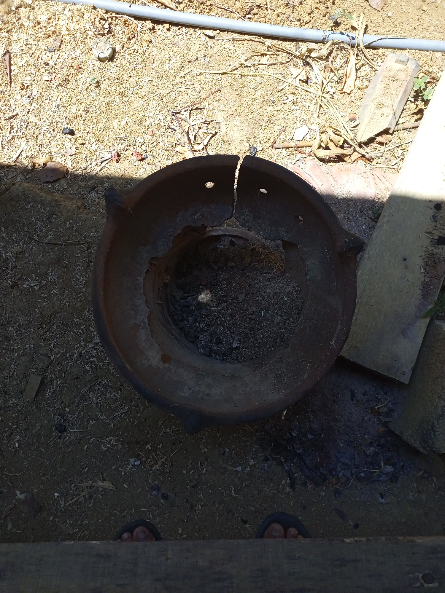 An overhead shot shows a broken, circular clay pot, likely a traditional stove, lying on the ground in Jatipurno, Indonesia. The pot is dark brown, cracked, and filled with ash and debris. It is surrounded by sandy soil, scattered leaves, and twigs. A gray pipe runs horizontally across the top of the frame, and a weathered wooden plank lies to the right of the pot. At the bottom of the frame, two bare feet in dark sandals are visible, peeking over a wooden surface. The scene appears to be outdoors, possibly in a yard or a rural area.