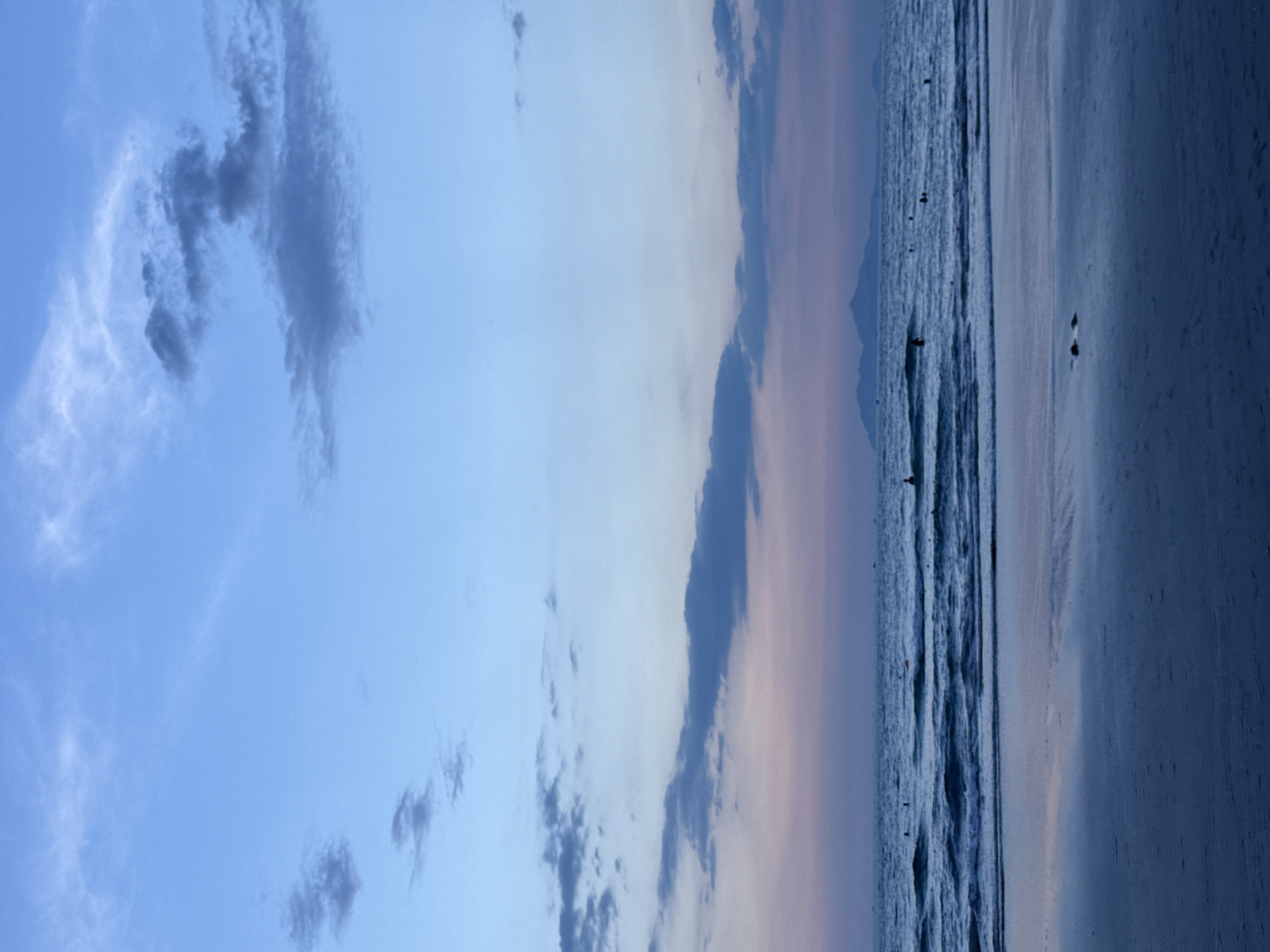 This serene landscape image, rotated 90 degrees clockwise from its natural orientation, depicts a tranquil beach scene at either dawn or dusk in Đà Nẵng, Vietnam. The sky dominates the upper portion of the frame, showcasing a gradient from a clear, bright blue at the top to lighter blues and soft, pale pink hues closer to the horizon, indicative of twilight. Scattered across the sky are various clouds, ranging from dark, textured formations to wispy, translucent streaks.
The horizon line extends across the middle of the image, separating the expansive sky from the ocean and coastline. In the far distance, a faint, dark silhouette of mountains or hills can be seen against the soft sky, suggesting a coastal topography. The ocean, appearing calm with gentle waves breaking near the shore, reflects the muted colors of the sky, creating a deep blue-grey tone.
Several small, dark figures are visible in the ocean, scattered throughout the mid-ground. These individuals appear to be enjoying the water, possibly swimming, wading, or surfing, adding a sense of human presence to the vast natural setting. The wet sandy beach, reflecting the ambient light, occupies the foreground.
The overall atmosphere of the image is peaceful and contemplative, capturing a quiet moment on the coast. There is no visible text within the image.