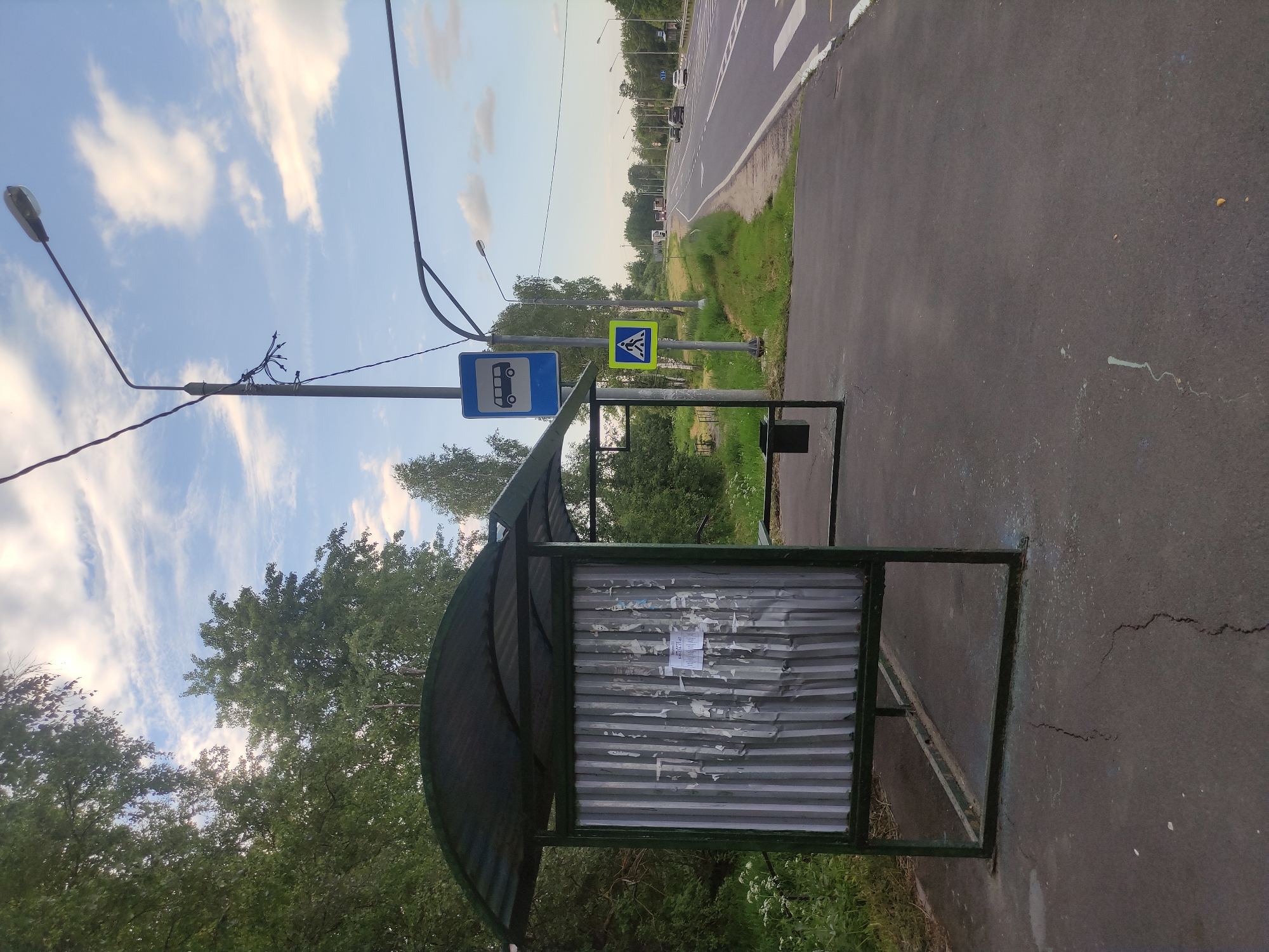The image depicts an outdoor scene at a bus stop in Kopylovo, Russia. A green-framed bus shelter with a corrugated metal back is positioned on an asphalt surface. A black trash receptacle is situated to the right of the shelter. Road signs are visible: a blue sign with a white bus icon and a yellow square sign with a white pedestrian icon. The bus stop is adjacent to a two-lane road with a pedestrian crossing marked by white stripes. Vehicles are present on the road in the distance. Trees and greenery surround the area, and the sky is partly cloudy. The overall environment appears to be a suburban or rural setting.