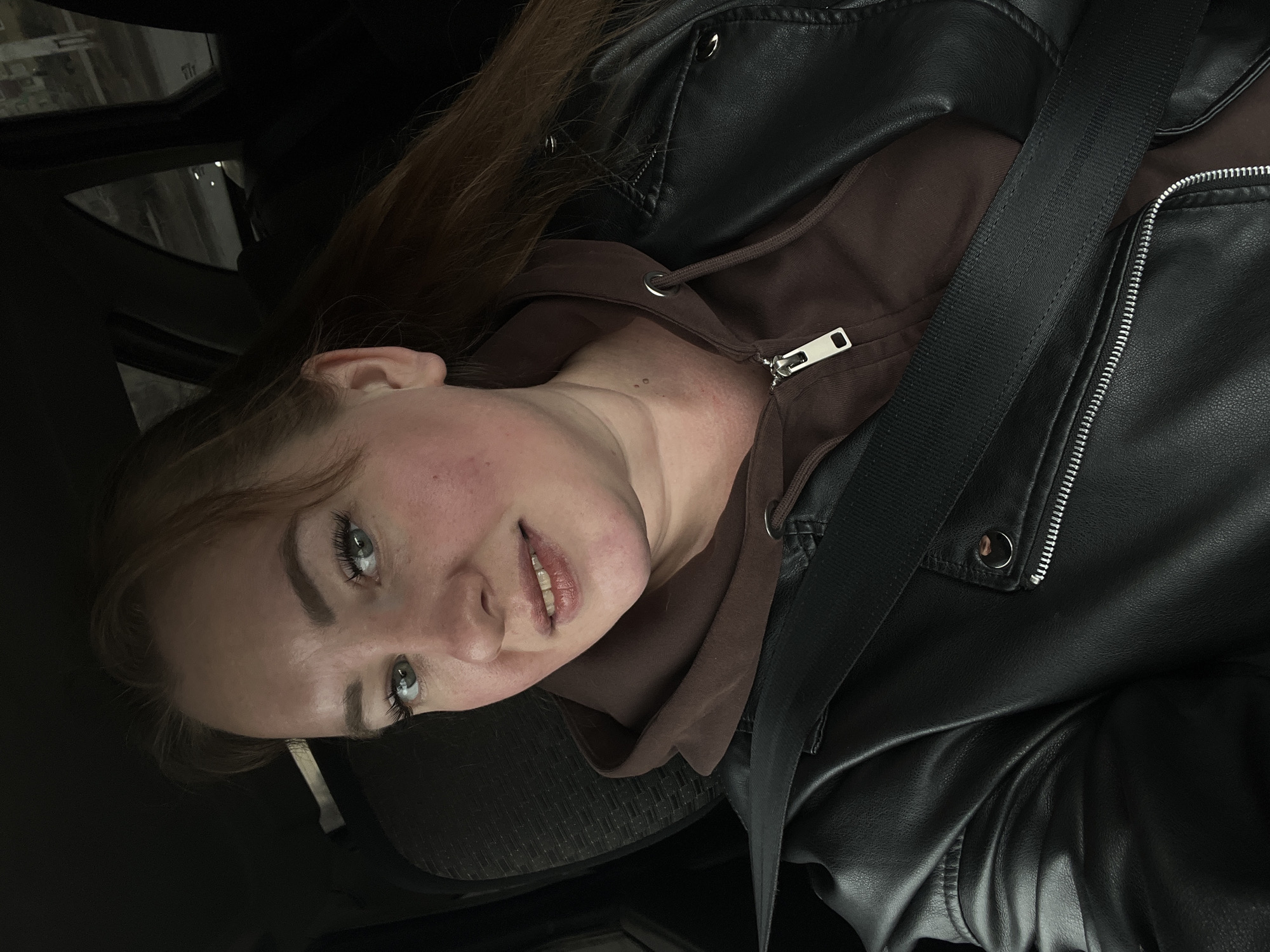 A young woman is photographed from a low angle inside a vehicle. She is wearing a brown hooded sweatshirt and a black leather jacket, secured with a black seatbelt. Her hair is reddish-brown, and her eyes are light blue or green. The background is dark and out of focus, but glimpses of the vehicle's interior and the urban environment can be seen through the windows. It is difficult to determine with certainty that the location is Chaikovsky, Russia without further information.