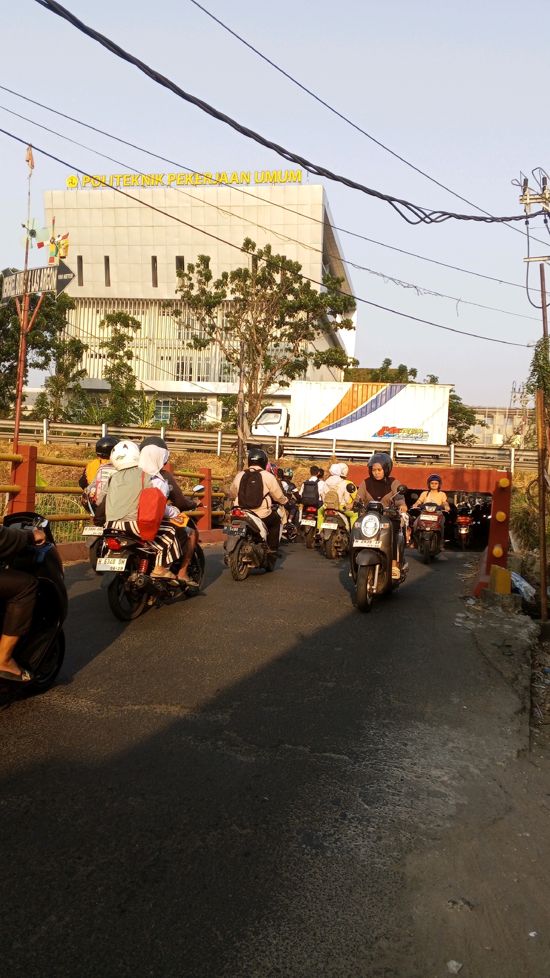 In Semarang, Indonesia, a traffic scene unfolds on a sunny day, likely in the afternoon given the long shadows. The image captures a busy street with multiple motorbikes, the primary mode of transportation. The motorbikes are moving in a queue, suggesting a traffic jam or a controlled intersection. Several riders and passengers are visible, many wearing helmets for safety, some in casual attire, and others with headscarves. One motorbike carries three people, one of whom has a red bag on their back.
Dominating the background is a modern, multi-story building with a light-colored facade, adorned with the prominent yellow lettering "POLITEKNIK PEKERJAAN UMUM" (Public Works Polytechnic). A large white truck with a blue and orange stripe logo is parked or moving behind a barrier, possibly related to the construction or operation of the institution.
The foreground and midground are occupied by the asphalt road, which shows signs of wear and tear, with some patches of lighter color indicating repairs or different material. A simple metal barrier with wooden posts runs along the side of the road. Trees with sparse foliage are present, indicating a potentially dry season. The sky is clear and bright blue, with several power lines crisscrossing the upper portion of the frame, a common sight in urban Indonesian landscapes. The overall impression is one of everyday urban life in Semarang.