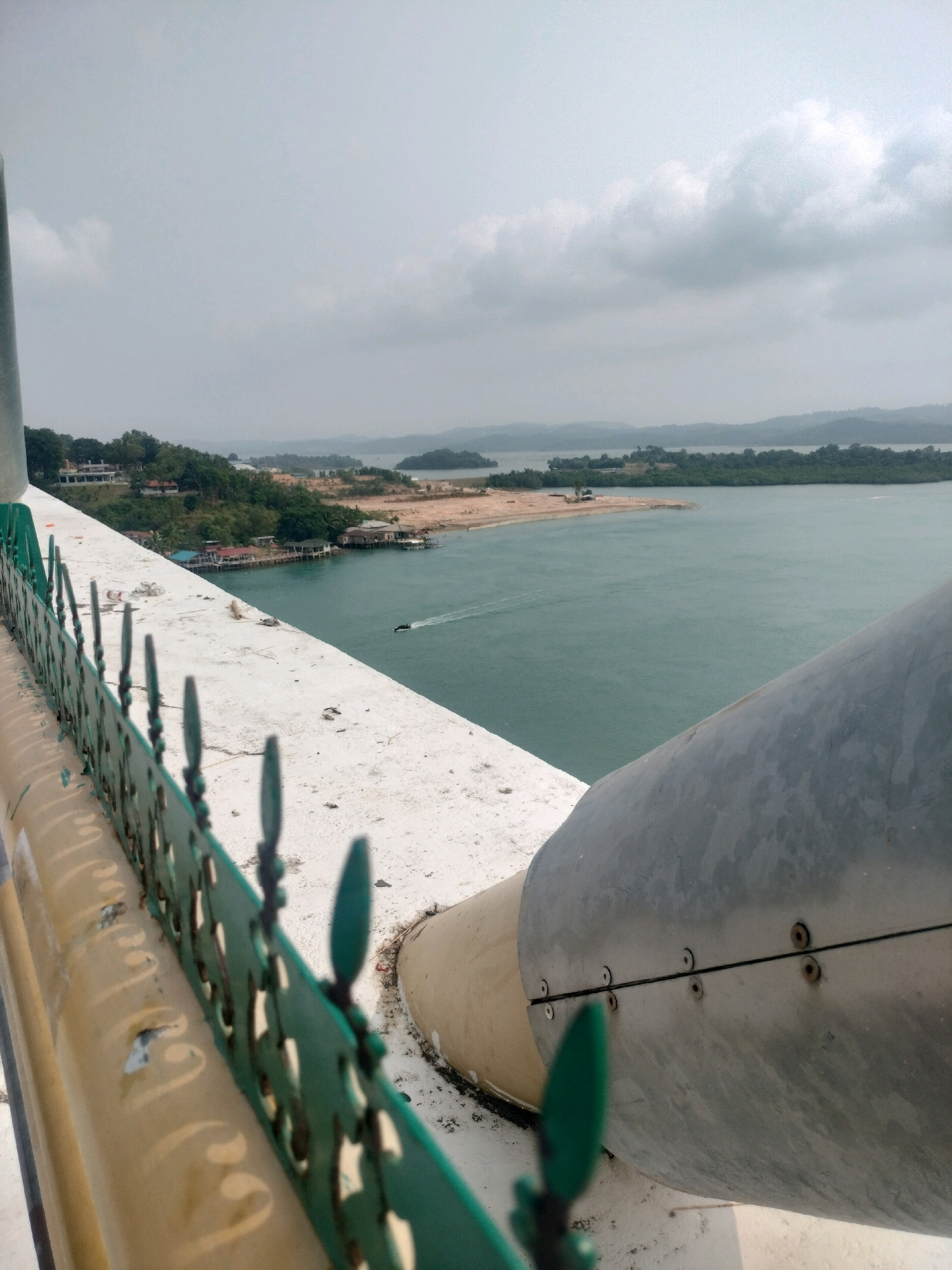 The image depicts a view from a structure overlooking a body of water. In the foreground, a metallic pipe or structural element is visible, attached with bolts. A green metal railing runs along the edge of the structure. Beyond the railing, a wide expanse of calm, greenish water stretches towards a distant, hilly coastline dotted with trees and some buildings. A small boat is seen creating a wake on the water. The sky is overcast with clouds. This scene is likely located in Batam, Indonesia, given the geographical features and architectural style.