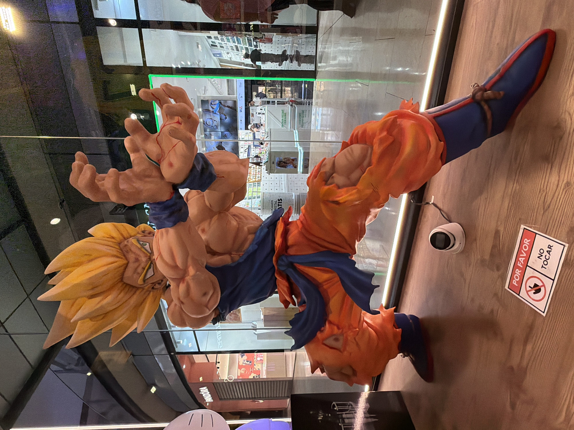 A large, full-body figure of the character Goku from the Dragon Ball franchise stands indoors. Goku is in a dynamic pose, wearing his signature orange and blue outfit. His spiky yellow hair is prominent. The figure is placed near a glass wall that reflects the interior of what appears to be a shopping mall or store. A security camera is mounted on the wall beside the figure, along with a sign in Spanish that reads "POR FAVOR NO TOCAR", indicating "Please Do Not Touch". The mall appears to be the ZAM Barcelona shopping mall in Barcelona, Spain. Visible shops include Pandora, Zam, Udon and Lacasa De Las Carcasas.