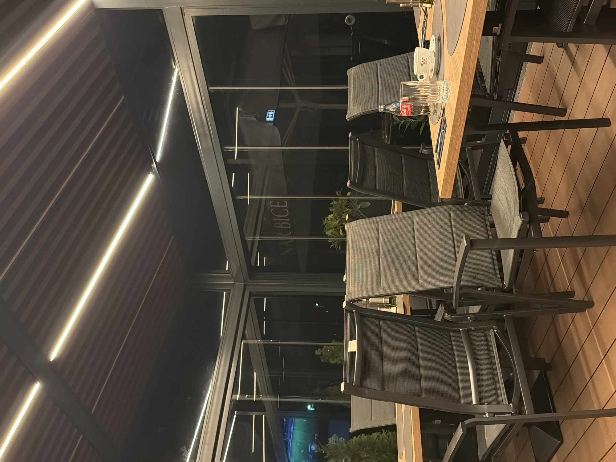 The image depicts an outdoor dining area, likely a restaurant patio, possibly located in Sofia, Bulgaria, as the signage "NAR BICE" is visible. There are several gray metal chairs arranged around a wooden-topped table. On the table, there is a cup, a bottle of Coca-Cola, and some dining utensils. The patio is enclosed with glass panels, offering a view of the surrounding environment, including greenery and lights. The roof structure is visible above, featuring linear lighting. The flooring is wooden. No people are visible in the frame.