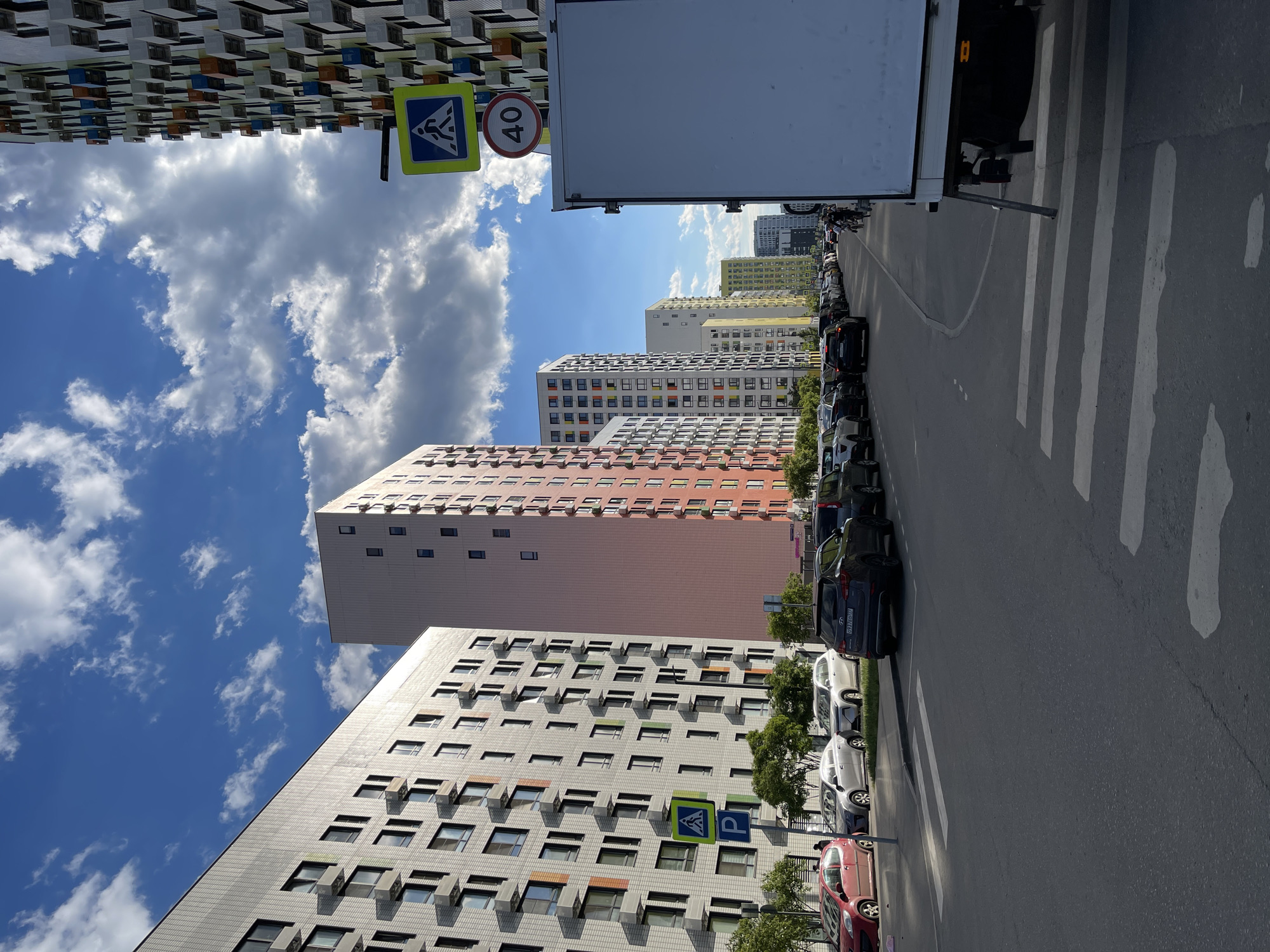 This image captures a vibrant daytime street scene in a modern residential area of Kommunarka, Russia. The view looks down a paved road, which gently curves to the left, with tall, multi-story apartment buildings lining both sides.
In the foreground, a wide pedestrian crossing is marked by thick white stripes on the dark asphalt road. A large white truck with a dark cab is partially visible on the right side of the road, facing away from the viewer. Behind the truck, a row of various cars is neatly parked along the roadside. Small trees are scattered between the parked cars and the buildings on the right.
The buildings display contemporary architecture, featuring repetitive patterns of windows and balconies. Their facades exhibit a range of colors; some are white or grey, while others have warm orange, yellow, or red tones, with occasional colorful accents around windows. On the far left, a distinct building features a complex, checkered pattern of grey, black, and colored (orange, blue, green) cube-like elements.
The sky is a bright blue with scattered white, fluffy cumulus clouds, indicating fair weather. Sunlight casts visible shadows.
Several signs are present: a yellow-bordered blue square sign with a white pictogram of a walking person (pedestrian crossing sign) is visible on both sides of the street. Next to one of these signs on the left, a circular sign with a red border displays "40", indicating a speed limit. Another blue rectangular sign with a white "P" (parking sign) is visible near the grey building on the right. Partially visible Russian license plates on parked cars (e.g., numbers "777" and "799") confirm the location context. No people are explicitly visible, suggesting a quiet moment in this bustling urban environment.