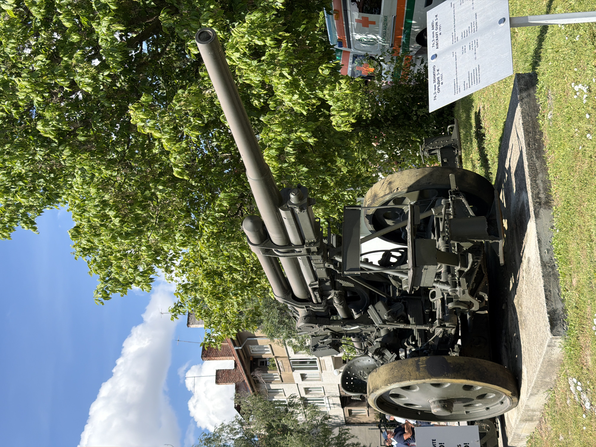 The image shows an anti-aircraft gun displayed outdoors, possibly as part of a museum exhibit. The gun is green and mounted on a wheeled carriage. A concrete base supports the gun. To the right of the gun is a sign with text in Bulgarian and English, detailing the specifications of the "76,2-mm ANTI-AIRCRAFT GUN 3-K M 1931". In the background, there is a building with multiple floors and windows, and lush green trees. An ambulance with a red cross emblem is partially visible behind the gun. The sky is blue with white clouds. There are a few people standing in the background, to the left of the gun, and a sign with text is also visible to the left. The location is identified as Sofia, Bulgaria.