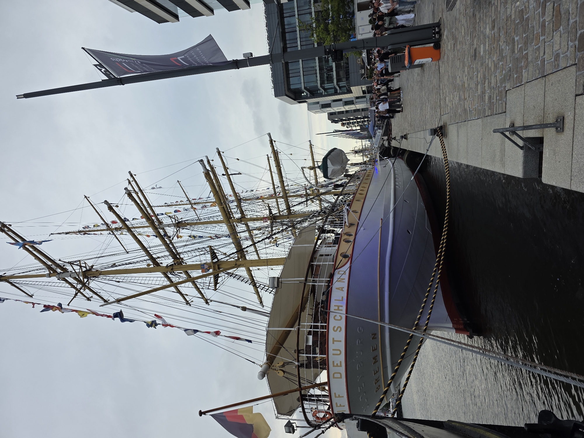 The image captures the sailing ship "Deutschland" docked at the Columbus Quay in Bremerhaven, Germany. The vessel, a historic tall ship, is adorned with numerous flags along its rigging, suggesting a festive occasion. The German flag flies prominently from its bow. The ship's name, "DEUTSCHLAND," is clearly visible on its hull, along with "OLDENBURG" and "BREMEN," indicating its homeport. The ship is moored to a cobblestone quay, where a crowd of people are gathered, some appearing to be tourists or visitors. Modern buildings, characteristic of a harbor or waterfront development, stand in the background. An orange waste bin is positioned on the quay. The sky is overcast, casting a muted light over the scene. The overall impression is that of a maritime event in a port city.