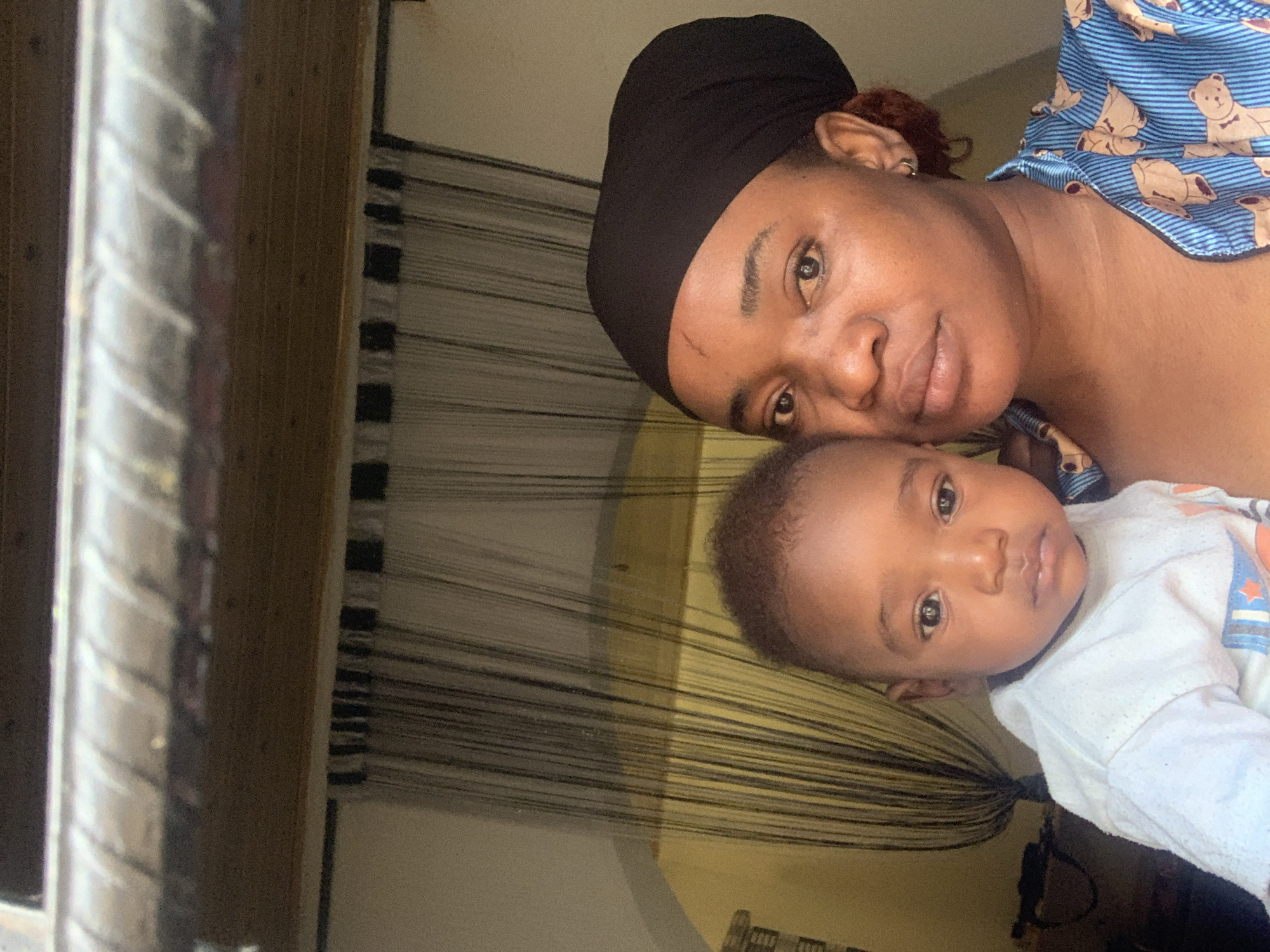 The image features an adult woman and an infant child in a close-up, horizontally oriented shot, likely taken indoors. The woman, with medium-dark skin, is positioned to the left of the frame, partially obscured by the baby, while the baby occupies the right side. Both subjects are looking directly at the camera.
The woman wears a dark headwrap and a blue and white striped top adorned with teddy bear patterns. Her expression is calm with a slight smile. A small scar is visible above her left eyebrow, and some reddish-brown hair can be seen near her ear.
The infant, also with medium-dark skin and dark hair, wears a white long-sleeved top with small, colorful patterns in blue and orange. The baby's expression is neutral and observant.
The background consists of a yellowish-brown surface, possibly a wall or bedding, and a series of thin, dark horizontal lines which might be part of a bed frame, window blinds, or room divider. To the far left, a silver-toned, ribbed metal bar or pole is partially visible. The lighting suggests natural daylight, creating a soft illumination on their faces. The overall mood is intimate and peaceful, capturing a moment of connection between the woman and child. No visible text is present in the image. The setting is consistent with an interior space, with the location context provided as Barakin Akawo, Nigeria.