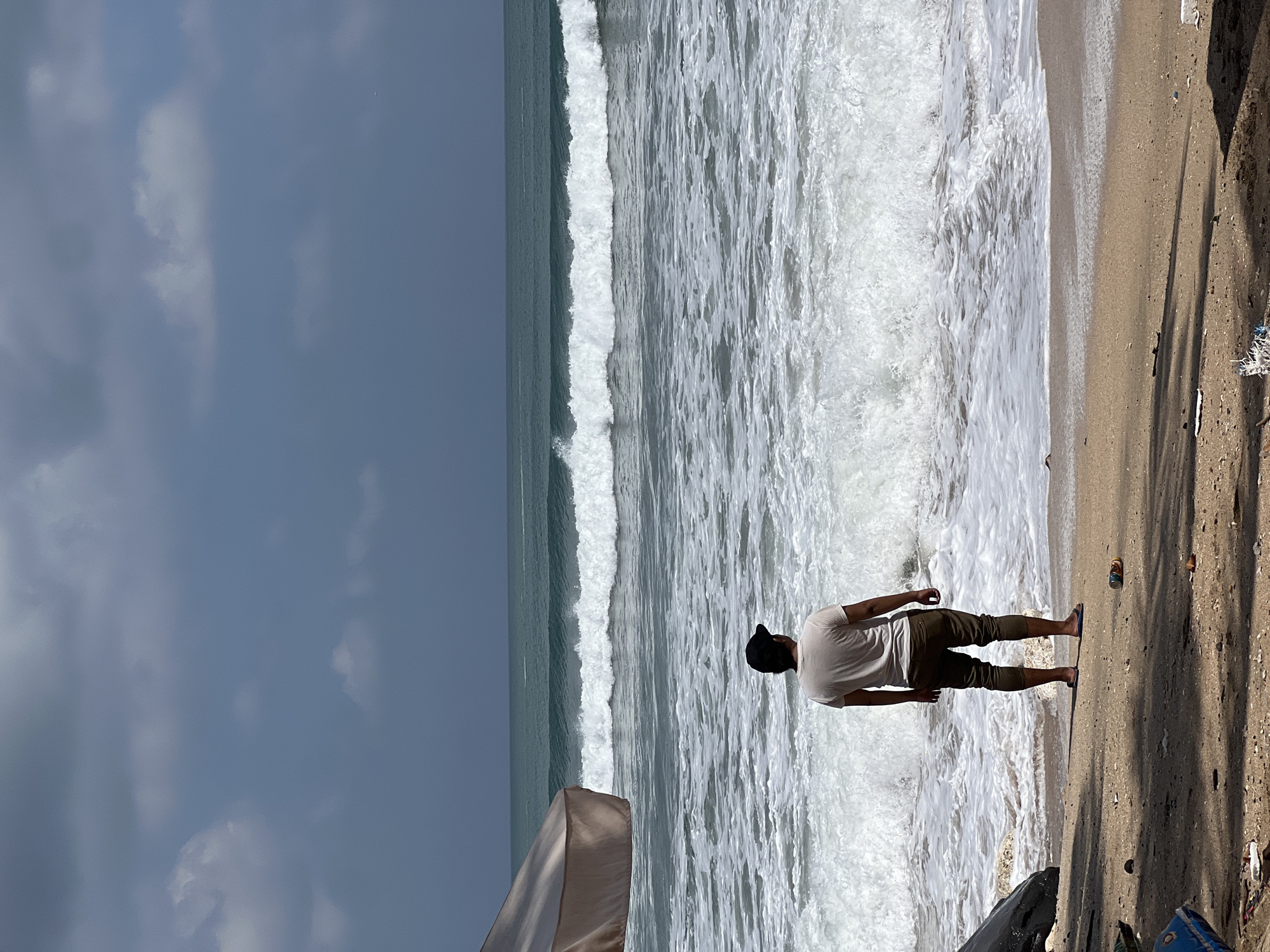 A man stands on a sandy beach, facing a turbulent ocean with large waves crashing ashore. The sky above is overcast and a dark gray. In the foreground, debris and shadows are visible on the sand. To the left of the image, a portion of a beach umbrella is present. The man is wearing a light-colored t-shirt, dark pants, and flip-flops, with a dark baseball cap on his head. He appears to be observing the sea. This scene is characteristic of Kuta, Indonesia, a popular beach destination known for its strong surf.