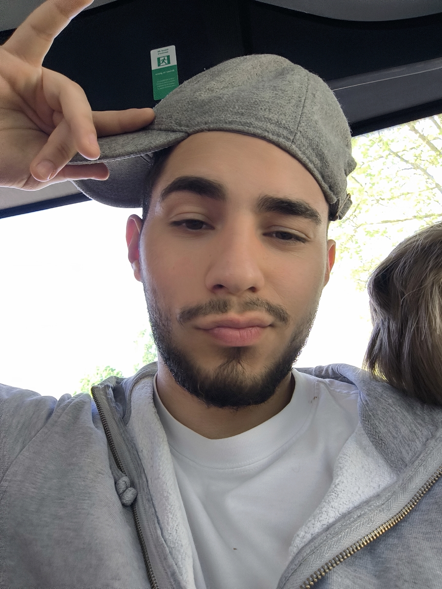 A selfie-style portrait shows a young man inside what appears to be a public transport vehicle, likely in Strasbourg, France. The man, with a beard and mustache, looks directly at the camera with a neutral expression and slightly pursed lips. He is wearing a light grey zipped hoodie over a white t-shirt and a light grey flat cap. His left hand is raised, forming a peace sign (V-sign) while also touching the brim of his cap.
To the right of the main subject, part of another person's head with light-colored hair is visible, suggesting they are seated next to him. The background outside the vehicle's window is brightly lit, revealing green foliage, indicating a sunny daytime setting.
Above the man's head, a green and white emergency exit sign is affixed to the ceiling. The sign displays the standard running person pictogram and includes text, which appears to be in French ("Sortie de secours") and German ("Ausgang"), consistent with the bilingual context of Strasbourg. The interior ceiling of the vehicle is dark. The image captures a casual, everyday moment, possibly during travel.