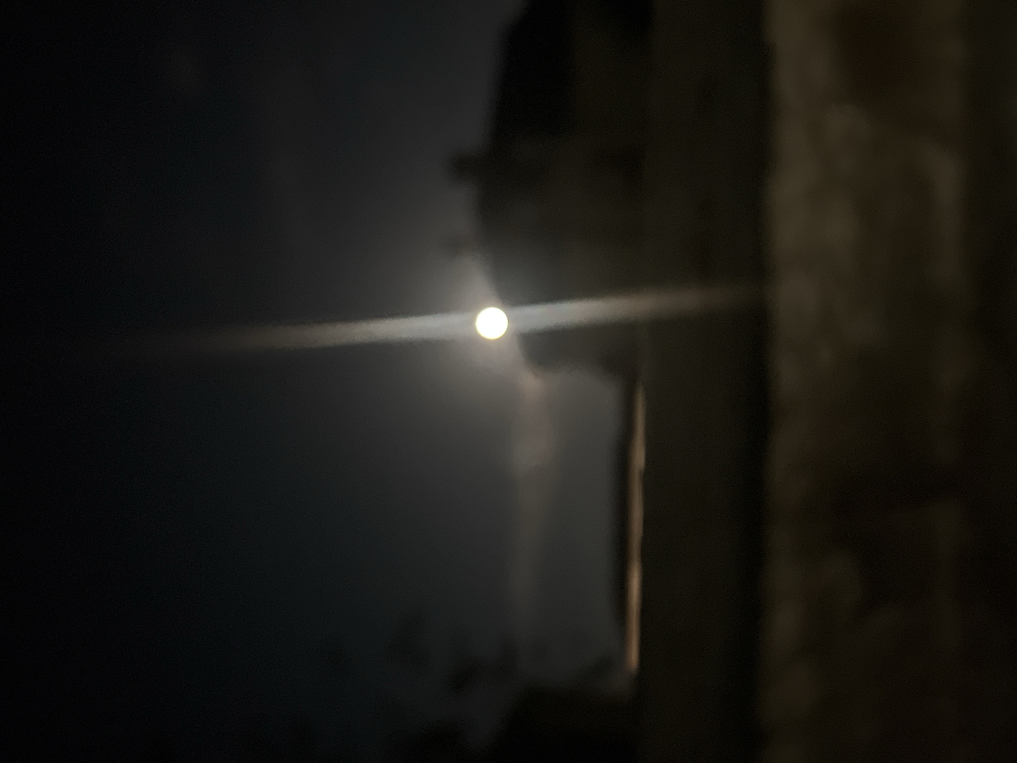 The image is a blurry, dark photograph taken at night. The moon is visible as a bright, out-of-focus orb in the upper center of the frame. A vertical streak of light extends downwards from the moon, obscuring some of the detail in the background. Silhouettes of buildings are faintly discernible behind the moon, and a horizontal band of lighter color, possibly a fence or a wall, runs across the middle ground. The foreground is dark and indistinct. The scene appears to be an outdoor setting, possibly a residential area. The image does not provide any specific details that would indicate the location within the city of Blagoevgrad, Bulgaria.