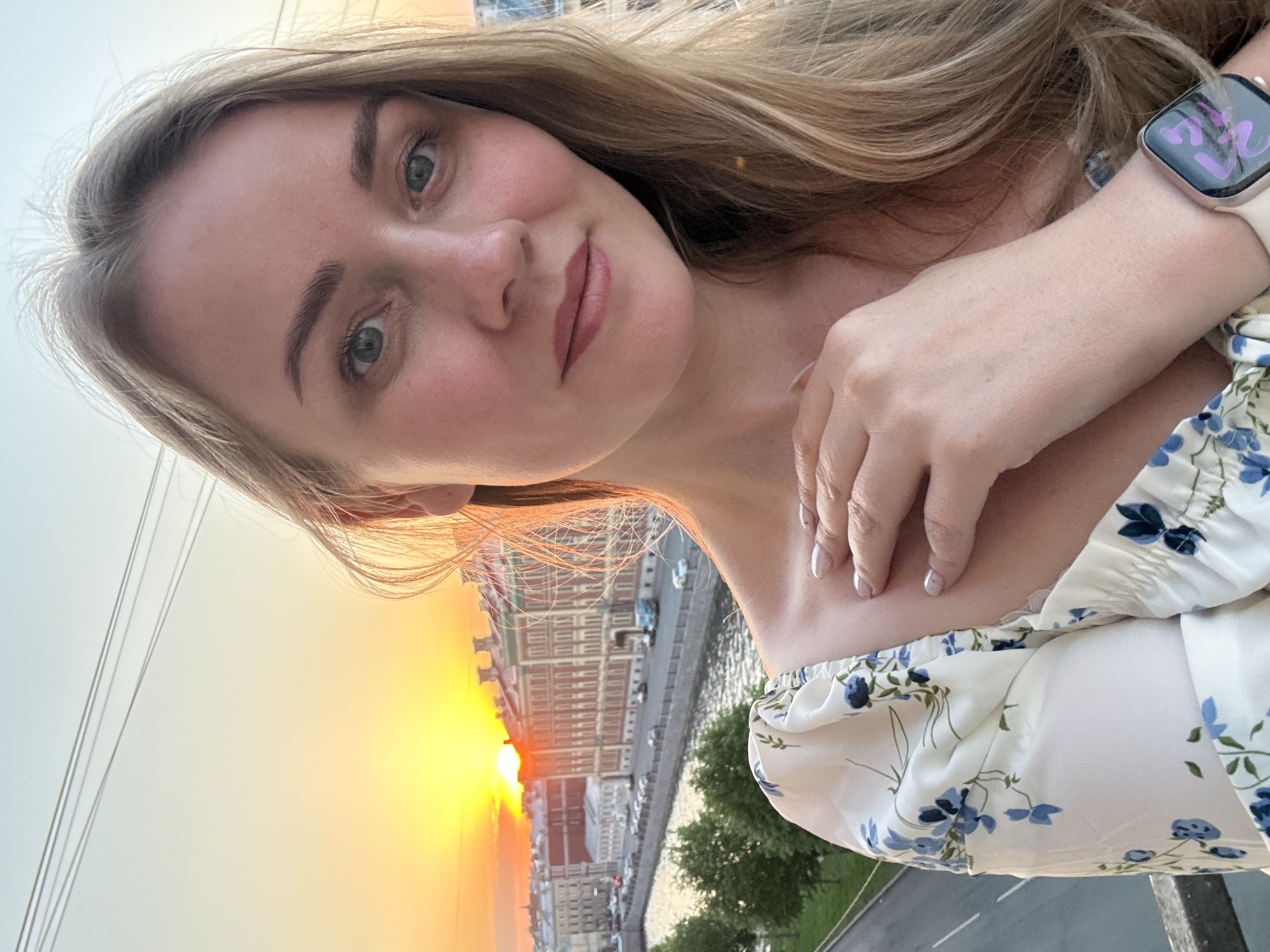 A selfie captures a fair-skinned blonde woman with blue-green eyes, looking directly at the camera. She wears a white dress with a blue floral pattern, and an Apple Watch is visible on her left wrist, displaying a purple-hued screen. Her hand is positioned near her neck.
In the background, the city of Saint Petersburg, Russia, is visible, notably the Neva River and historic buildings illuminated by a sunset. The sun is positioned low on the horizon, casting a warm light. Power lines are visible in the upper left quadrant. Trees and a street line the river's edge.