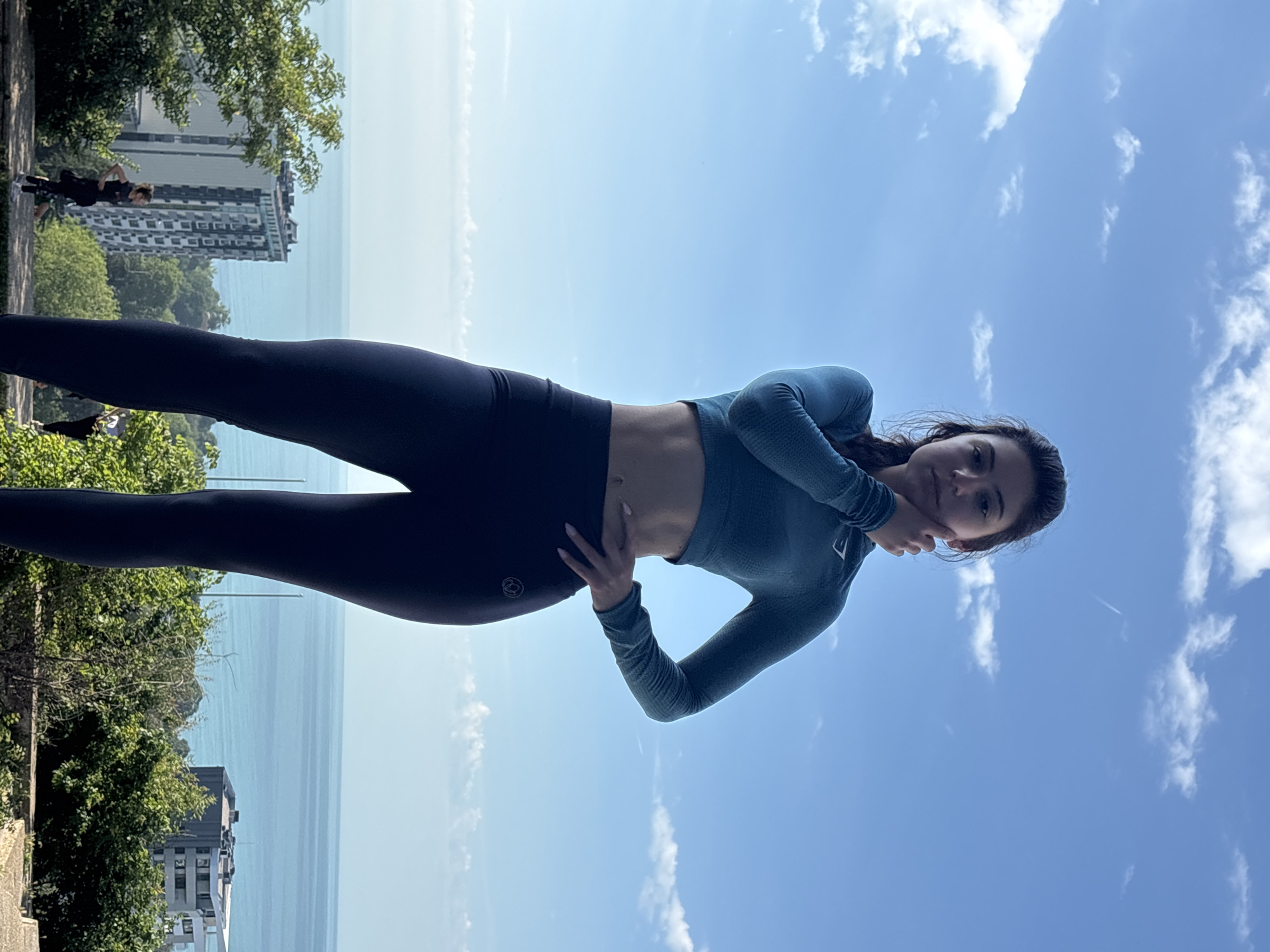 The original image, rotated 90 degrees counter-clockwise, depicts a young woman posing outdoors on a sunny day in what appears to be a coastal location, identified as Varna, Bulgaria.
The main subject is a young woman with dark hair, dark eyes, and fair skin, looking directly at the camera with a subtle expression. She is wearing a form-fitting, long-sleeved athletic top in a teal or blue color, and dark leggings. Her left hand is placed on her hip, and her right hand rests lightly on her chin. A brand logo, partially visible as "SMYRN" or similar, is on her right sleeve, and a circular logo featuring "22" is present on her left thigh.
The background is split between a vast, calm blue sea and a bright blue sky with scattered white clouds. The horizon line is clear and distinct. To the woman's left, abundant green foliage from trees and bushes forms a natural barrier, with several multi-story buildings visible in the mid-distance along the coastline. Farther back on the path, small, indistinct figures suggest other people are present.
The scene takes place during bright daylight, indicative of good weather. The woman is posing for a photograph. The combination of the sea, coastal buildings, and greenery aligns with the provided location context of Varna, Bulgaria.