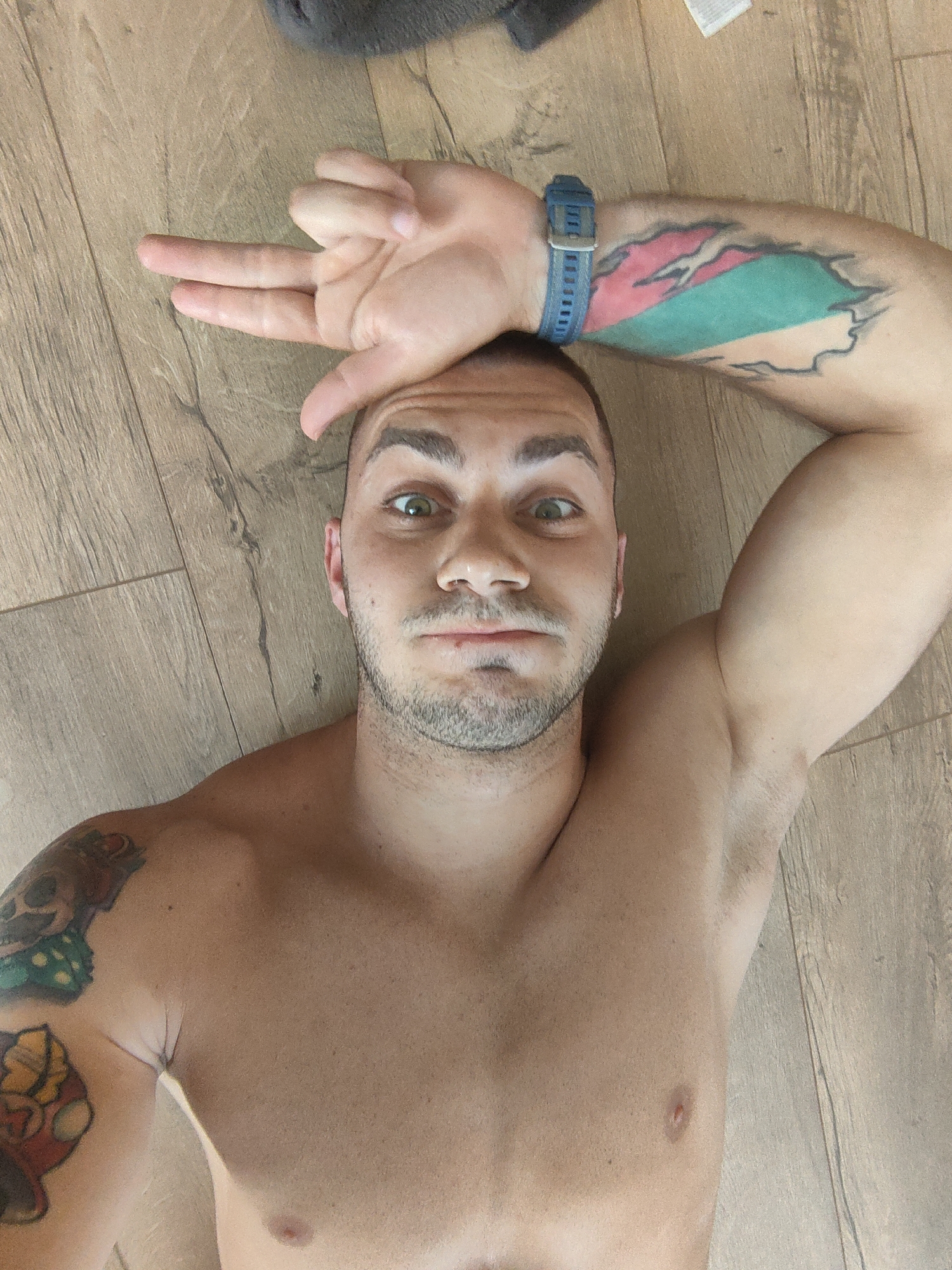 A shirtless man with a tattoo on his left arm and shoulder lies on a wooden floor, looking up at the camera. He is holding his right hand above his forehead, with two fingers extended. The man has short, dark hair and stubble. A blue watch is visible on his left wrist. The background is a wooden floor with visible grain patterns. The image is likely taken indoors. There is no information about the location in Varna, Bulgaria.