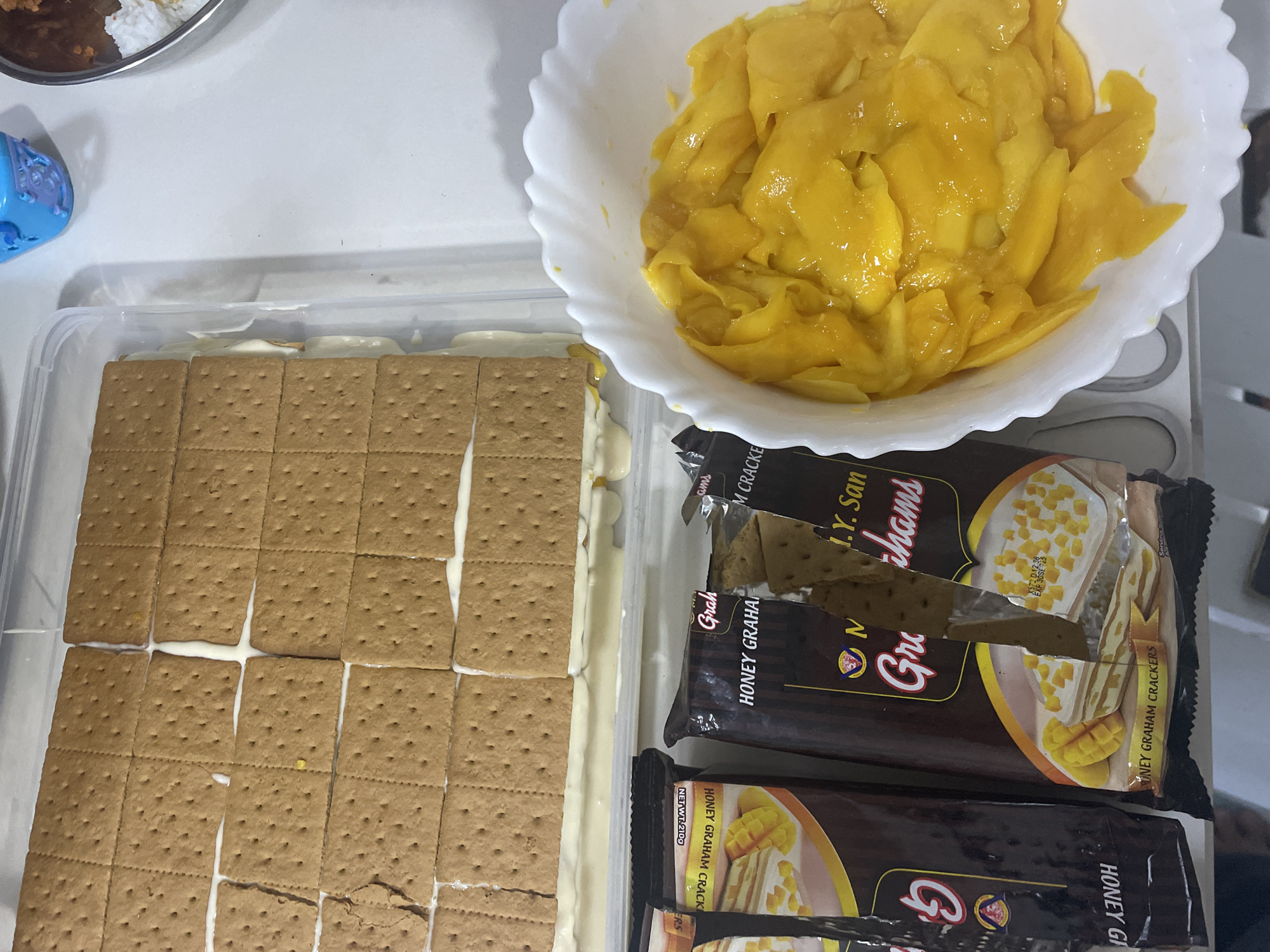 The image shows a partially assembled no-bake dessert, likely a mango graham cake, within a clear rectangular plastic container. The dessert consists of layers of graham crackers and a creamy filling. The graham crackers are arranged in a grid pattern. To the right of the container is a white, scalloped-edged bowl filled with sliced, ripe mangoes. In the foreground, two opened packages of "Honey Graham Crackers" are visible, with one package showing a cut-out revealing a picture of the finished dessert with mangoes. The setting appears to be a kitchen or dining area, with a white countertop. The presence of mangoes strongly suggests a location in the Philippines, where mangoes are abundant and widely used in desserts. Specifically, Davao City is renowned as the "King of Fruits" hub in the Philippines, known for its high-quality mangoes, making it a plausible location for the ingredients and preparation of such a dessert.