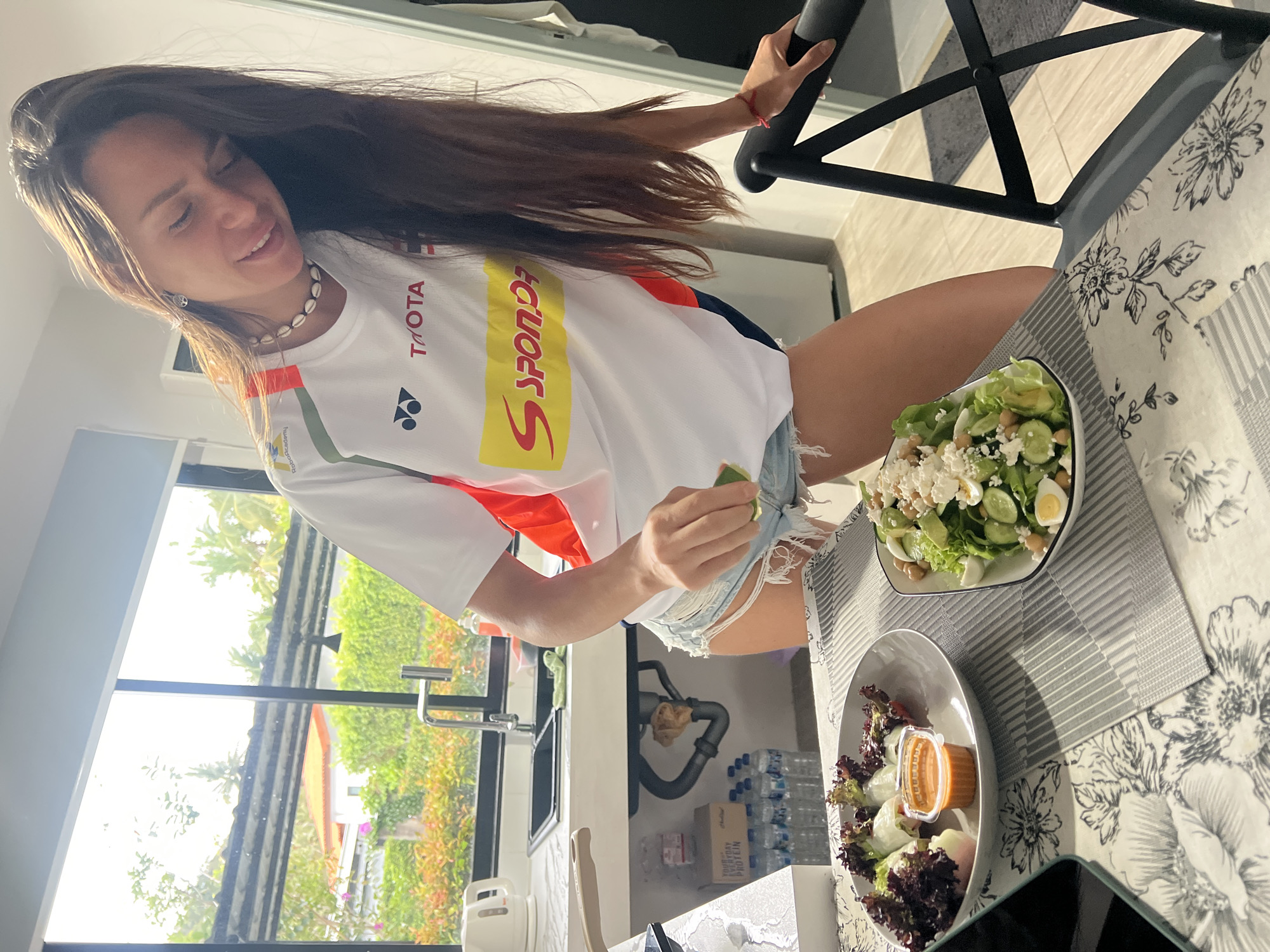 A woman with long brown hair wearing a white shirt and denim shorts is sitting at a table in what appears to be the kitchen of a house in บ้านบางรักษ์, Thailand. She is holding a piece of lime. Two plates of salad are on the table. The kitchen features a window with a view of tropical vegetation. Water bottles are visible under the sink.