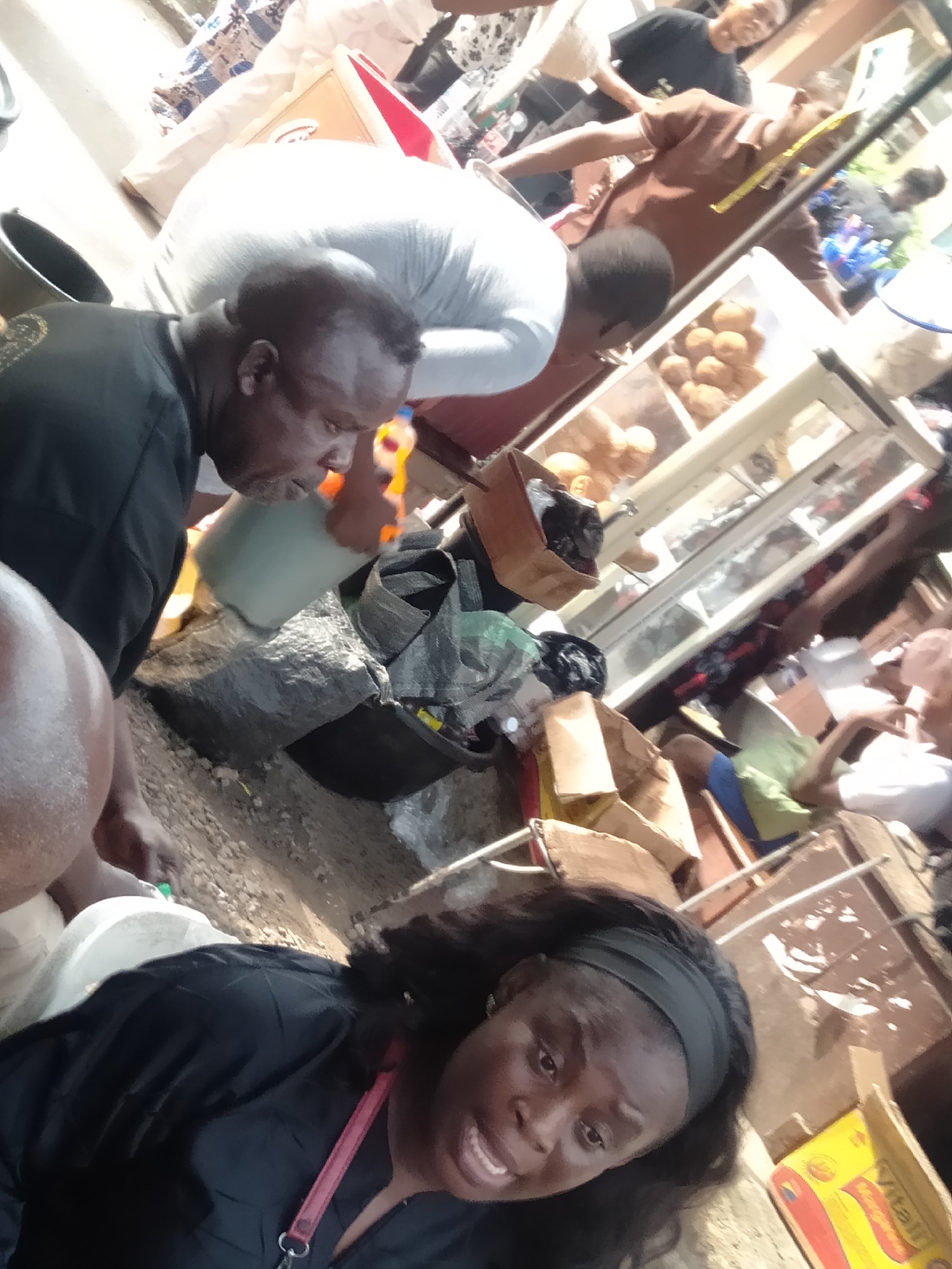 The image depicts a bustling street scene in what appears to be Lagos, Nigeria, evident from the yellow "Vital" margarine box and the general chaotic and informal setting.
In the foreground, a woman with a black headband and a black shirt, with a pink strap or lanyard visible, looks directly at the camera with an open-mouthed expression. To the left and slightly behind her, a dark-skinned man in a black shirt is pouring something from a light-colored container into a dark bucket or bin. There are various boxes and debris surrounding him.
Behind these two, the scene transitions into a more cluttered market or roadside vending area. Another person in a grey top appears to be reaching into or working on something. To the right is a display case containing what looks like pastries or snacks. In the background, additional individuals are visible, dressed in casual attire, seemingly engaged in various activities. The environment is cluttered, reflecting the lively and informal nature of street commerce in a major city. The ground appears to be rough and uneven. The scene conveys a sense of activity, commerce, and everyday life.