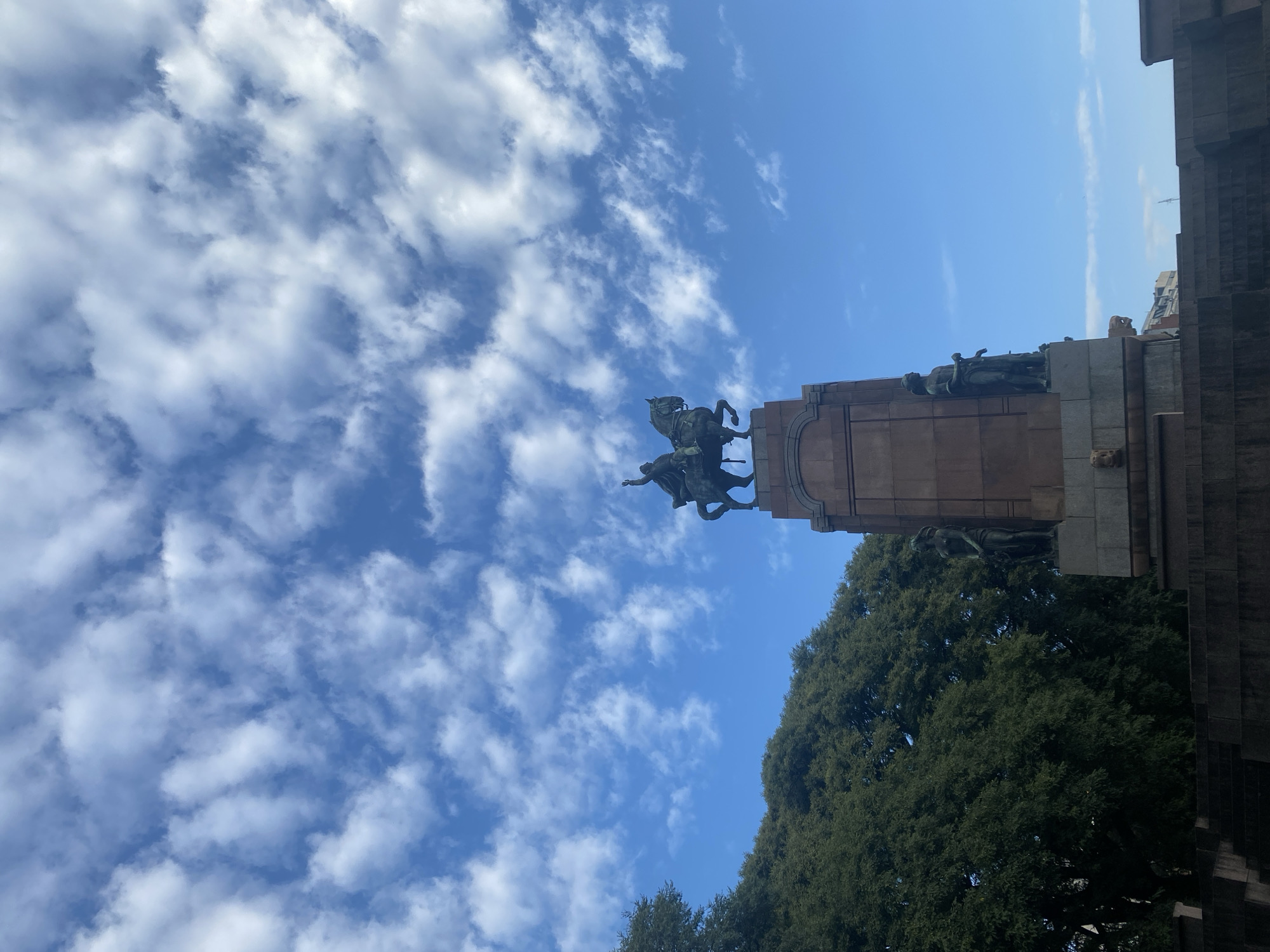 The image, initially rotated 90 degrees clockwise, depicts a grand, multi-tiered monument set against a vibrant blue sky with scattered white clouds. The central feature is a prominent equestrian statue crowning the monument, showing a figure on horseback in a dynamic, rearing pose. Below or alongside the horse, another sculpted human figure is visible, appearing to gesture or point. Several other human figures are integrated into the lower sections of the monument's stone structure.
Lush, dark green treetops are visible in the lower part of the image and to the right, suggesting the monument is located within a park or a tree-lined urban area. On the far right, part of a dark, angular building or structure can be seen, indicating an urban environment.
The bright lighting suggests it is daytime with clear, sunny weather. The low-angle perspective, looking upwards, emphasizes the impressive scale and intricate details of the monument against the expansive sky. No text is visible in the image. Given the context, this monument is located in Buenos Aires, Argentina.