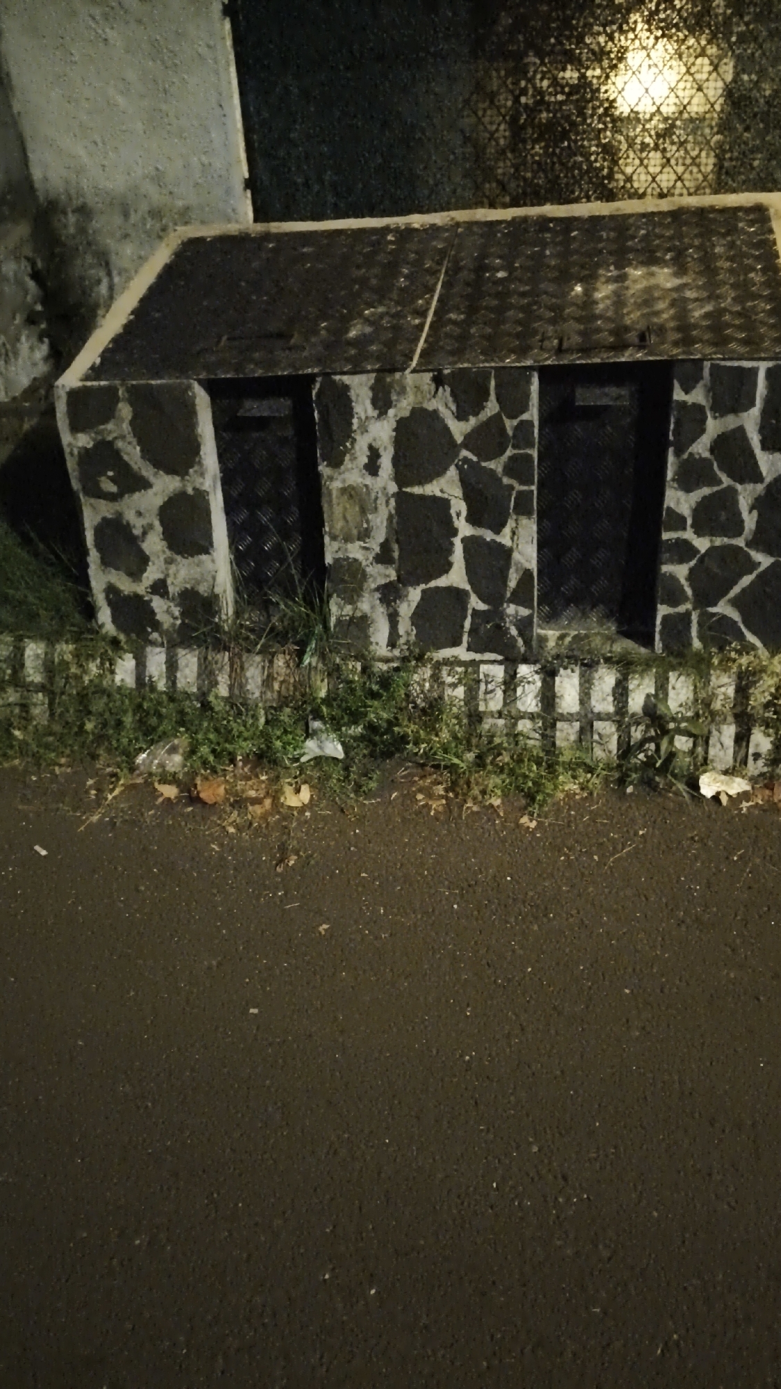 The image depicts an outdoor scene at night, likely in a city environment. In the foreground, a dark, asphalt surface, possibly a road or sidewalk, is visible. The midground features a low, rectangular structure constructed with irregular, dark stones. This structure has a textured, dark-colored roof and appears to have at least two openings or entrances, suggested by darker rectangular sections within the stone facade. A small patch of green vegetation, including grass and some scattered debris like leaves and plastic, grows along the base of this structure, separated from the asphalt by a low, white, grid-like barrier. In the background, a dark, textured wall or fence with a repeating pattern is present, with a bright, diffused light source visible behind it, casting a patterned reflection. The overall impression is of a utilitarian or infrastructural element within an urban setting. Based on the provided context, this scene is located in South Jakarta, Indonesia.